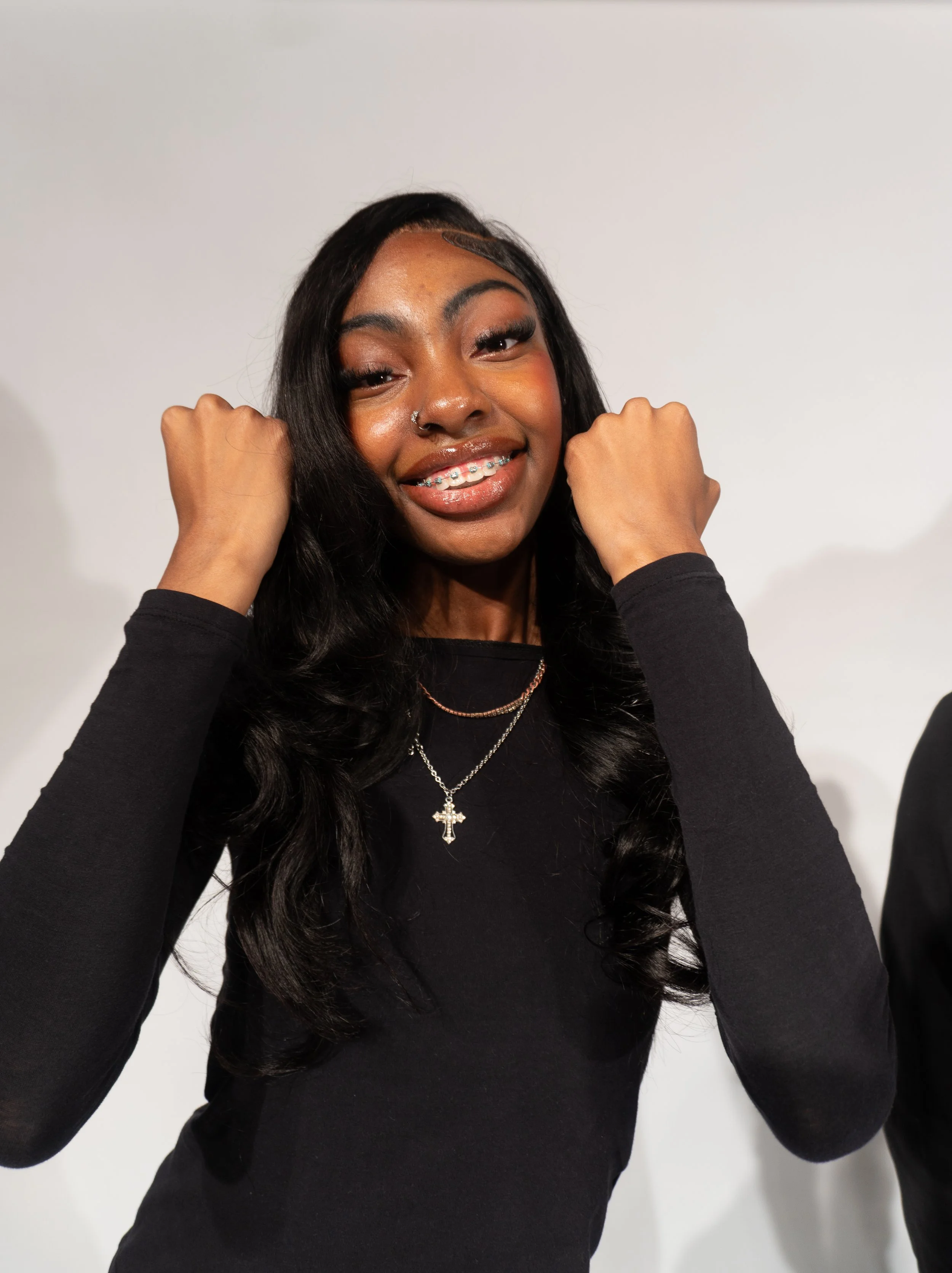 A young woman with long black hair, smiling, showing braces, wearing a black long sleeve shirt and necklaces, raising her fists near her face, standing against a plain white background.