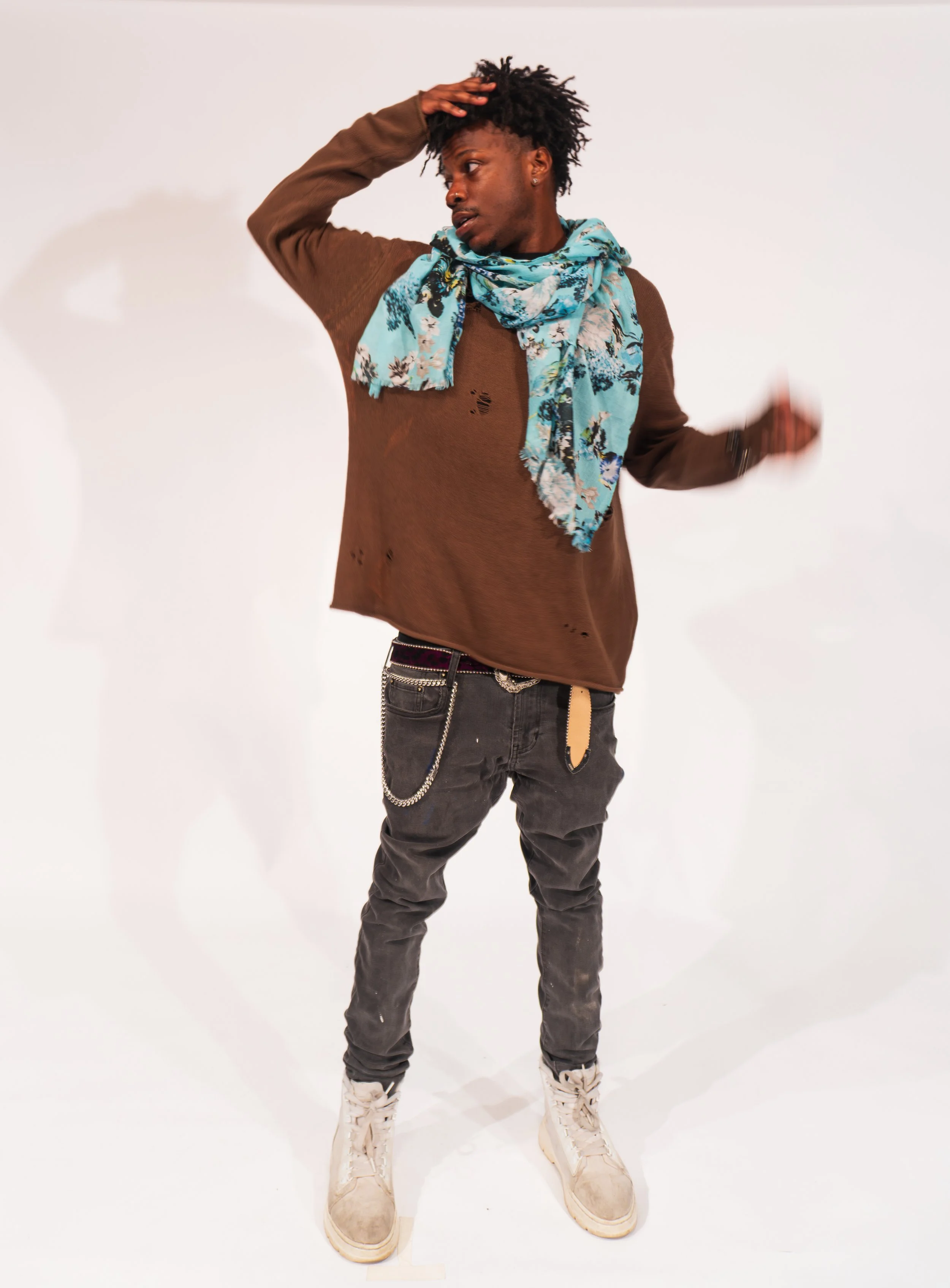 Young man with curly hairstyle wearing a distressed brown sweater, patterned blue scarf, black jeans with chains, and beige boots, standing against a white background.
