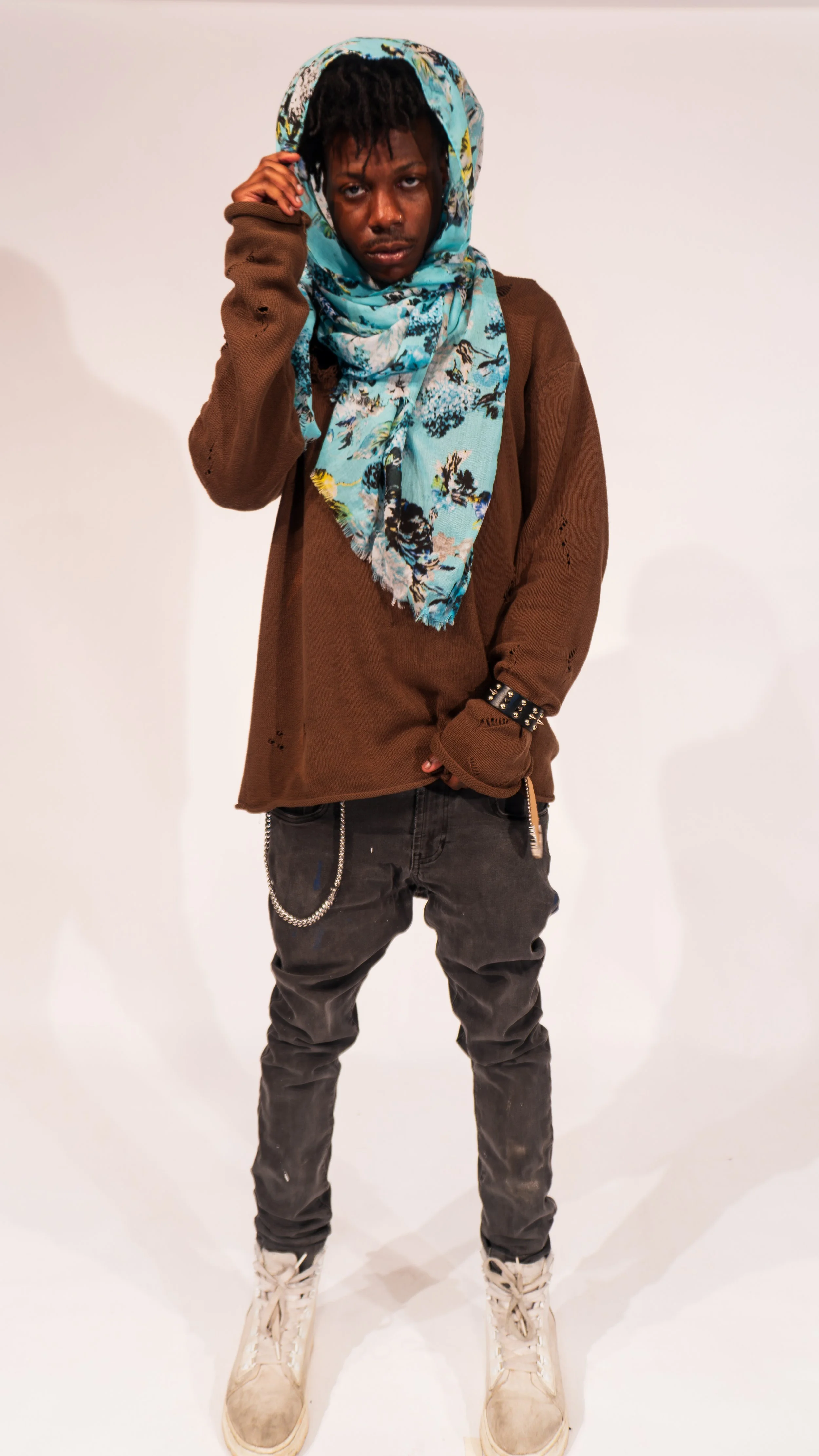 A young man with dreadlocks wearing a light blue floral scarf on his head, a brown distressed sweater, black jeans with a chain, and off-white sneakers, standing against a plain white background.