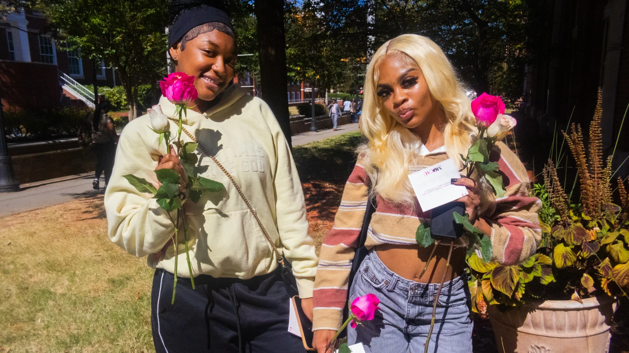 Two young women standing outdoors on a sunny day, holding pink and white roses, with trees and a park in the background.