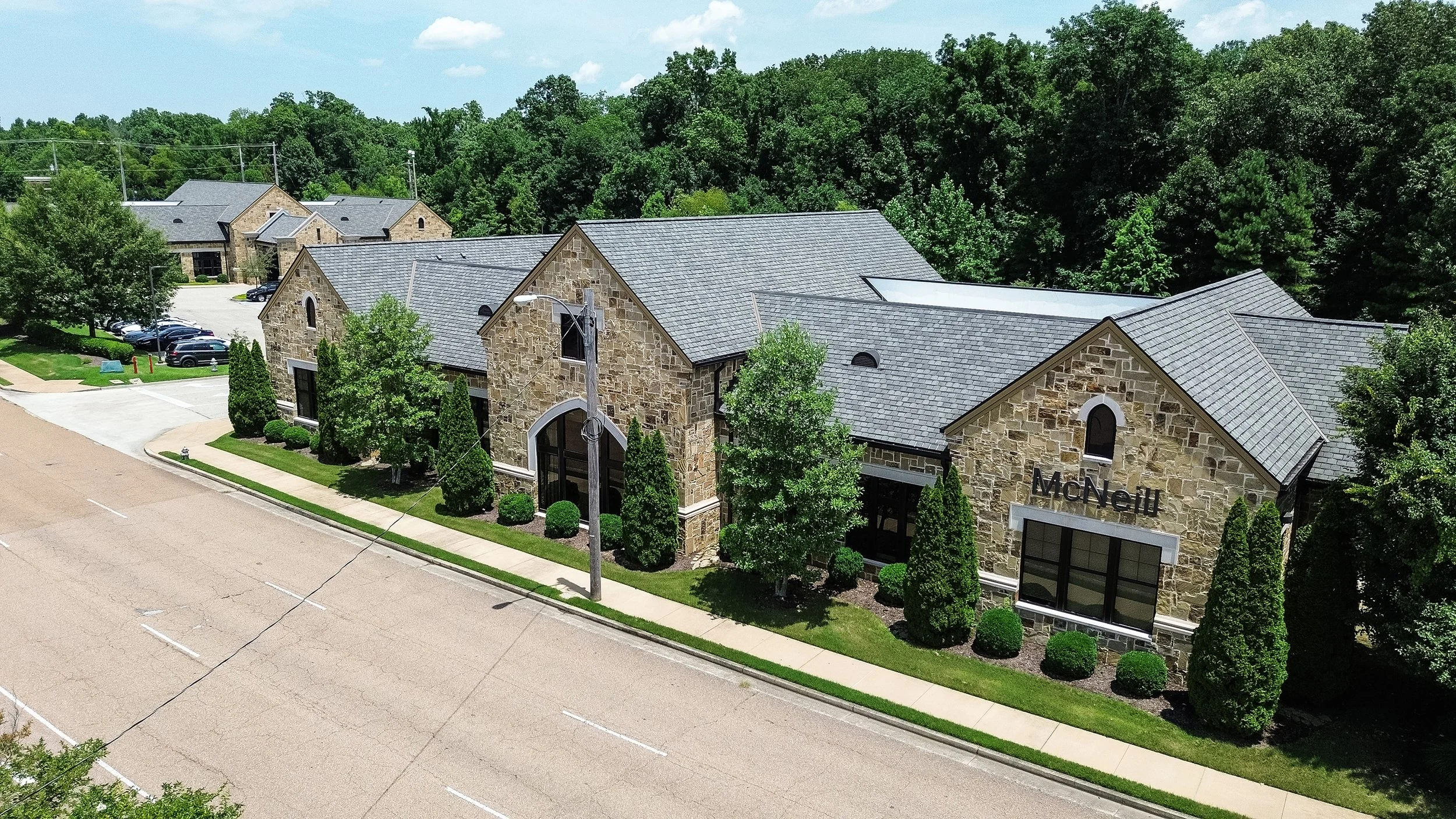A stone building with the name 'McNell' on it, featuring large arched windows, surrounded by green trees and shrubs, with a parking lot and a wooded area in the background in Germantown, TN.