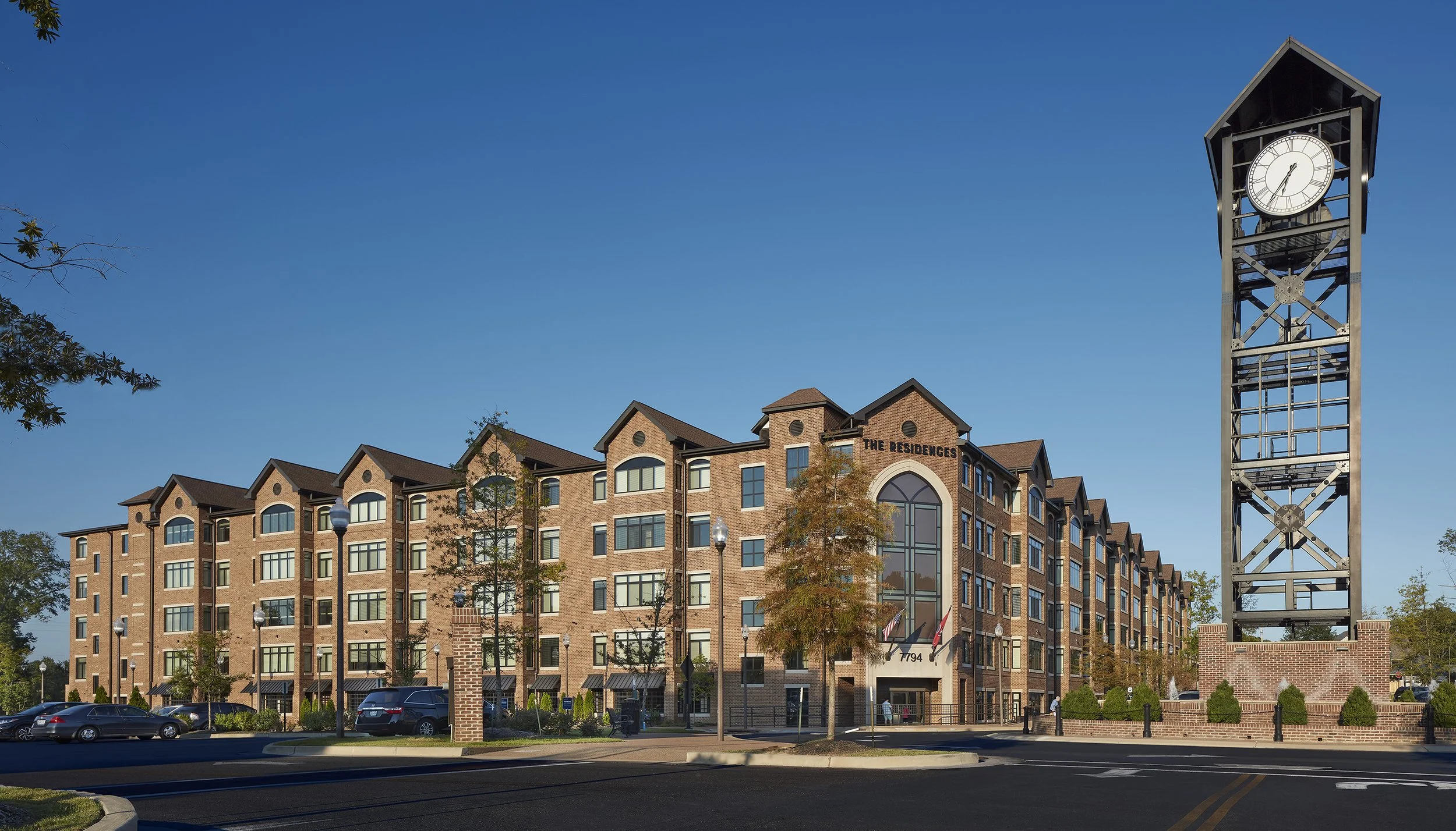 A multi-story brick apartment building named 'The Residences' in Germantown, TN. A tall clock tower with an analog clock is situated to the right of the building, and there are parking spaces with cars, trees, and street lamps in the foreground.