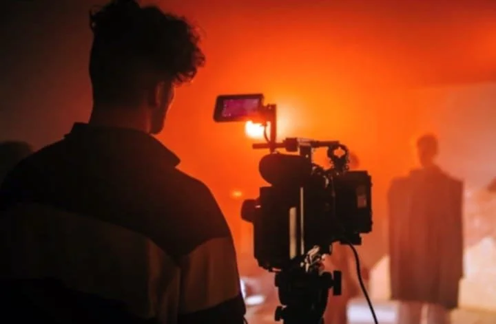A person filming with a professional camera on a tripod in a dark, smoky environment with orange lighting, and a person standing in the background.