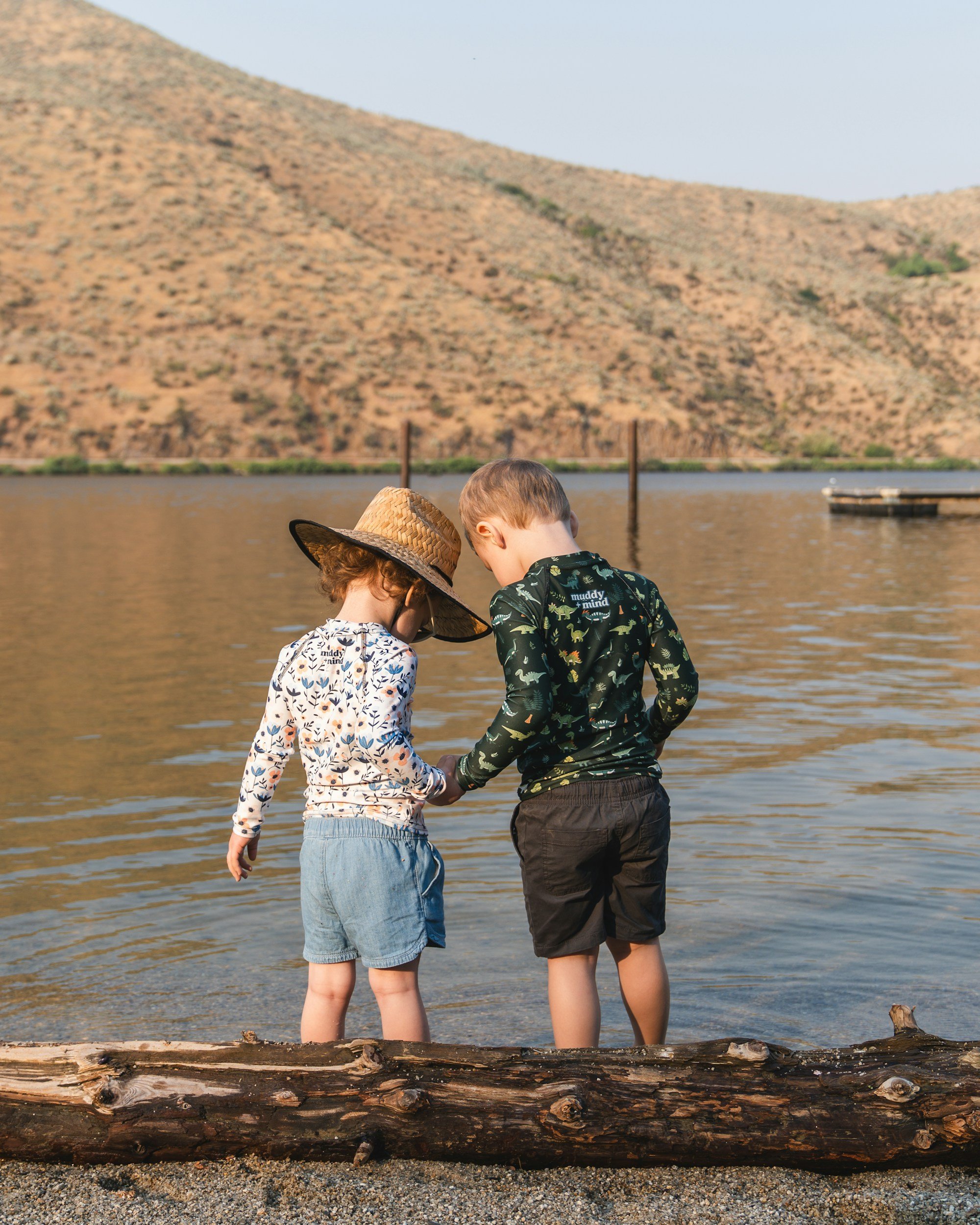 Two children are standing at the edge of a lake, holding hands, with mountains in the background. One child wears a wide-brimmed hat and a long-sleeve shirt with a floral pattern. The other wears a long-sleeve shirt with dinosaur print and shorts.