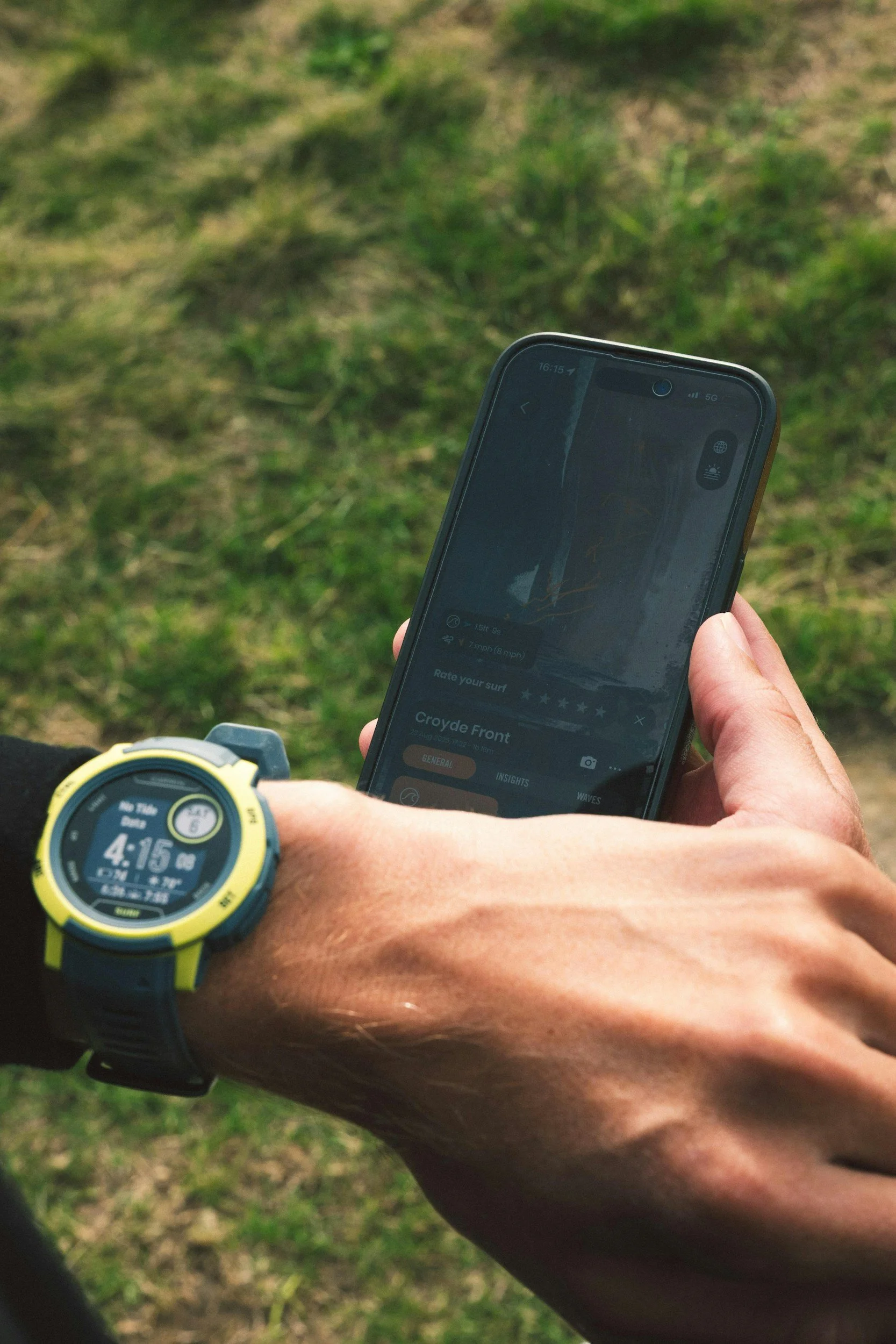 A person wearing a yellow and black sports watch is holding a smartphone displaying a surfing app on a grassy outdoor setting.