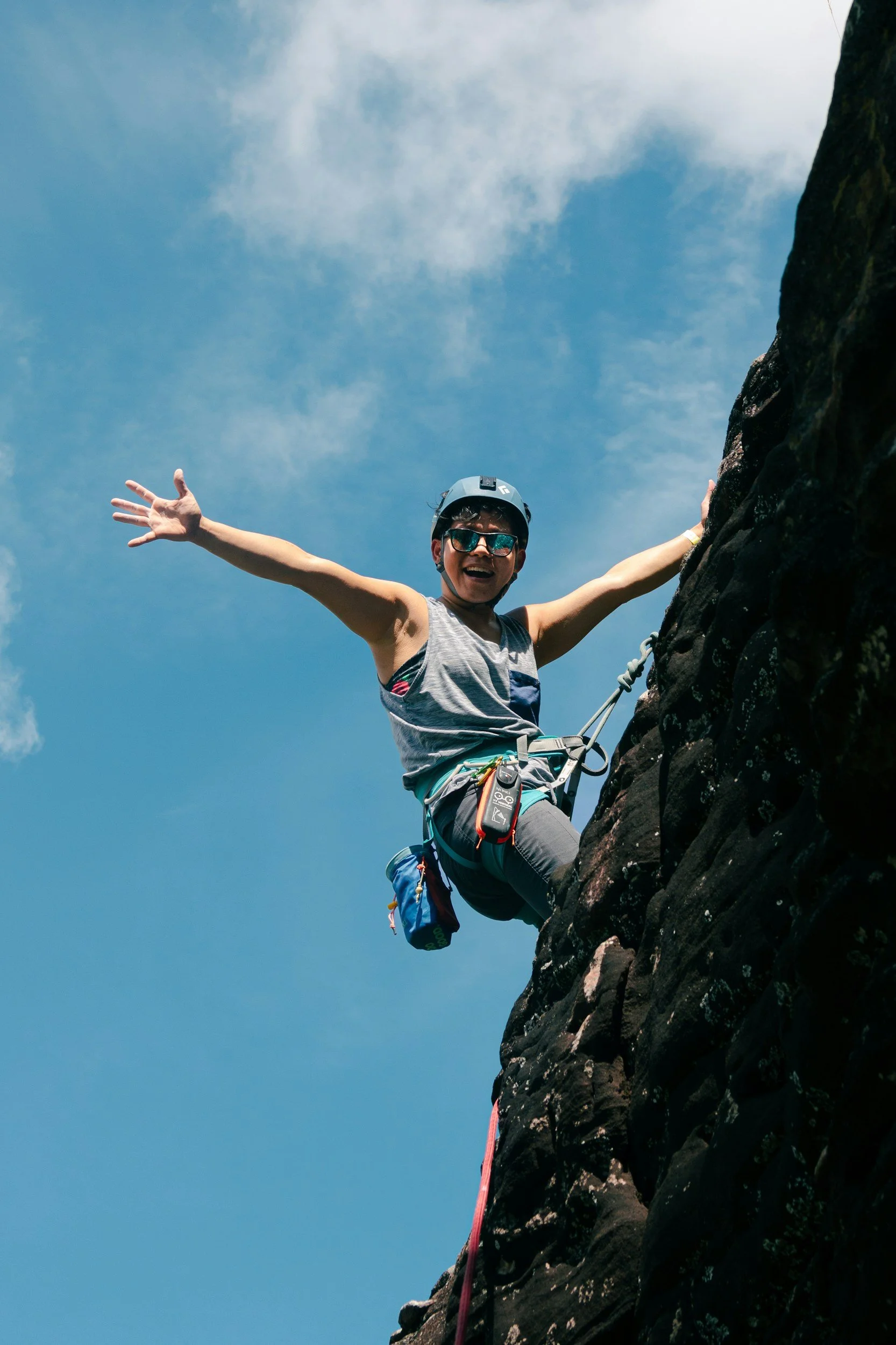 A person wearing a helmet and sunglasses is rock climbing on a steep cliff, smiling and extending arms outward against a blue sky with some clouds.