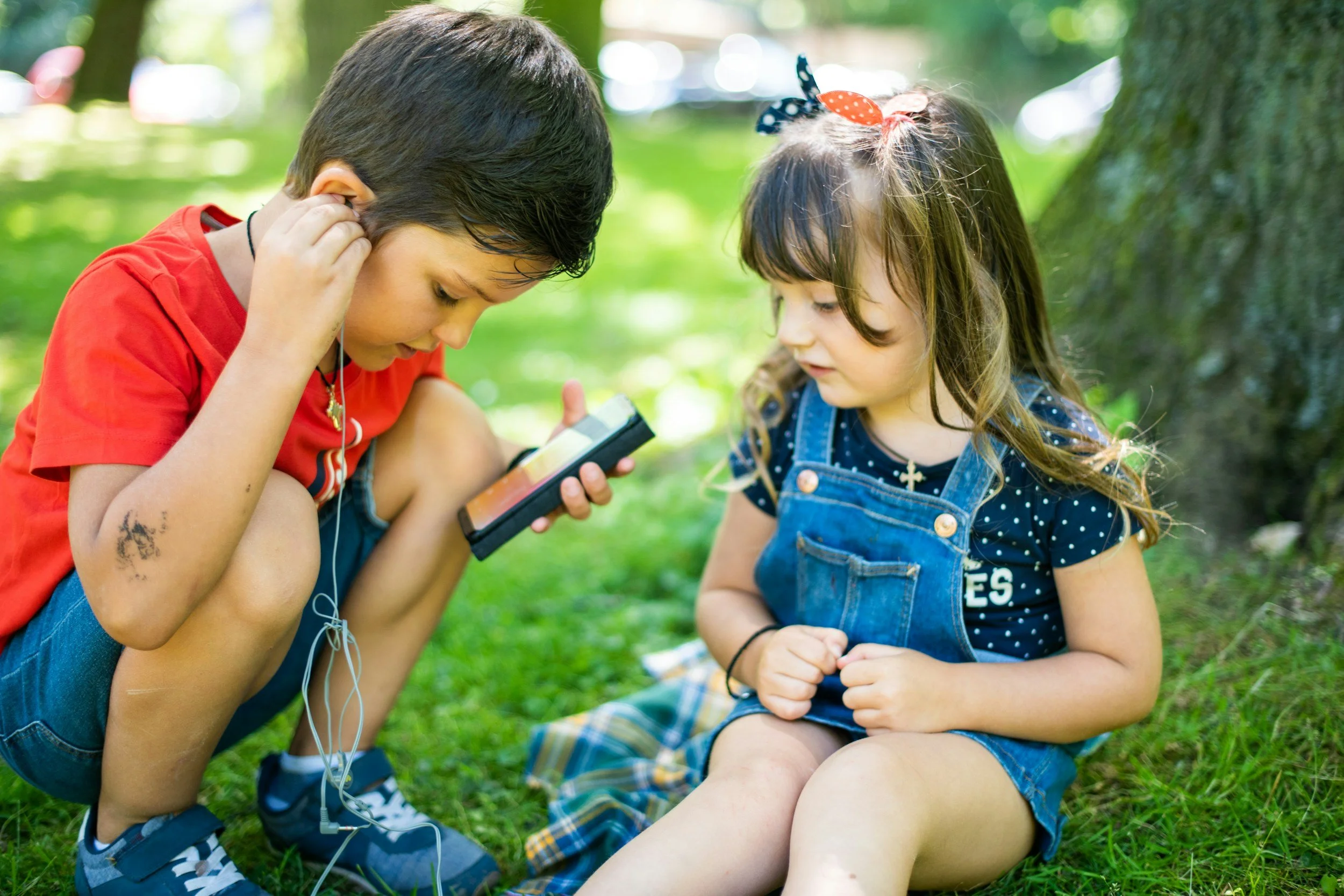 Two children, a boy and a girl, are sitting on the grass outside near a tree. The boy is wearing a red shirt and shorts, holding a device, and listening with earphones. The girl is wearing a blue shirt and denim overalls, looking at the boy and the device.