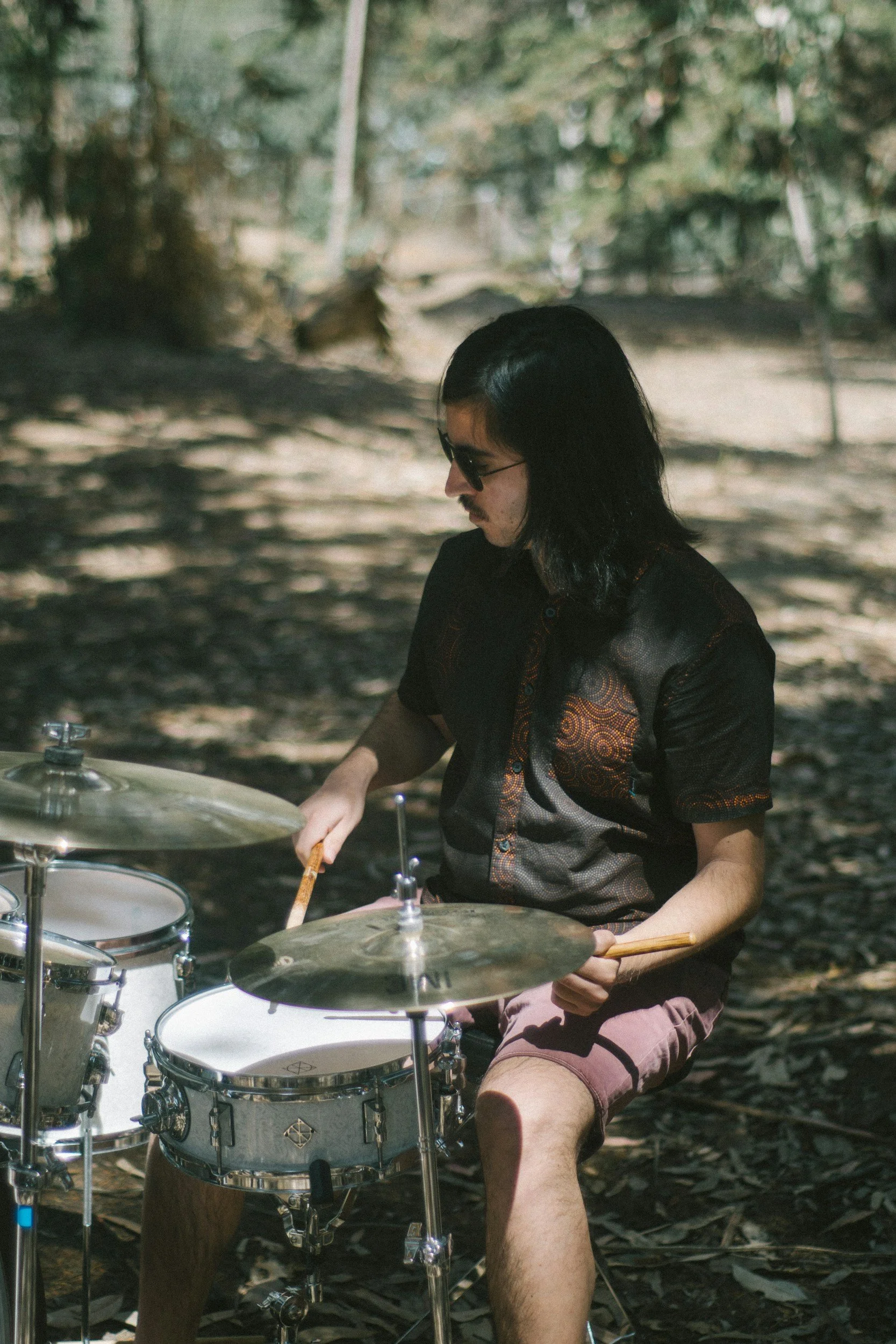 A man with long black hair, sunglasses, and a mustache sitting outdoors playing a drum set surrounded by trees and shadows.