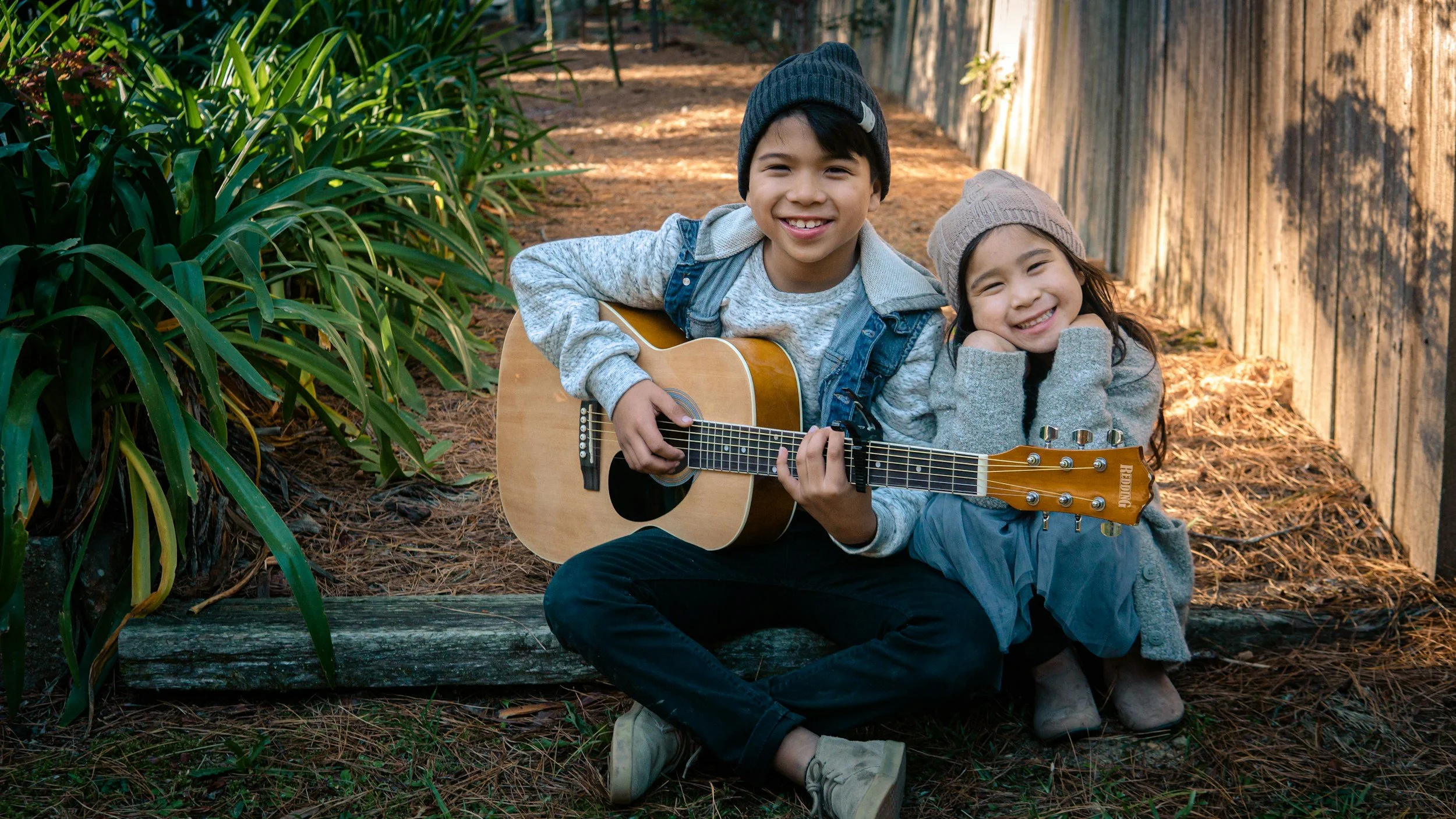 A boy playing guitar and a girl smiling while sitting outdoors by a wooden fence and green plants.