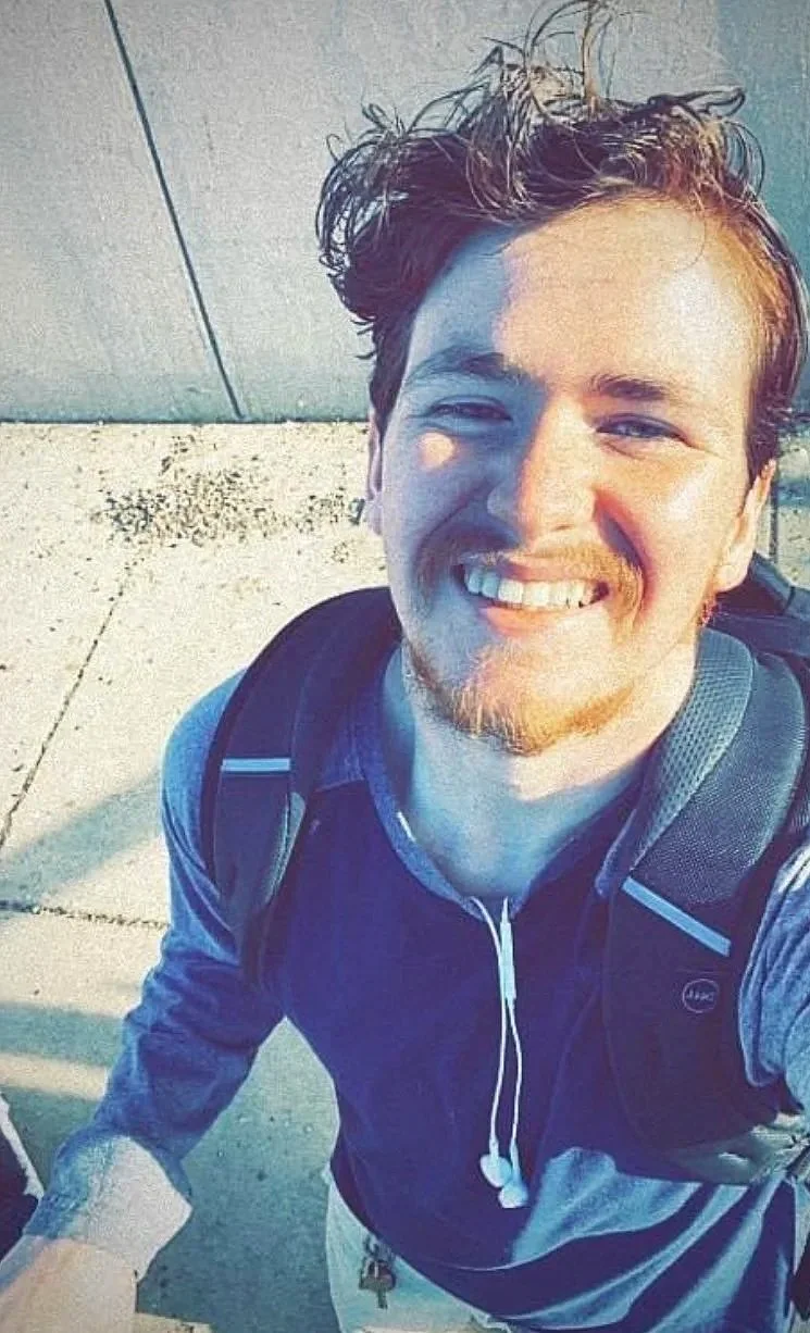A young man smiling outdoors, wearing a backpack, a blue and gray long-sleeve shirt, and earphones, with sunlight highlighting his face.