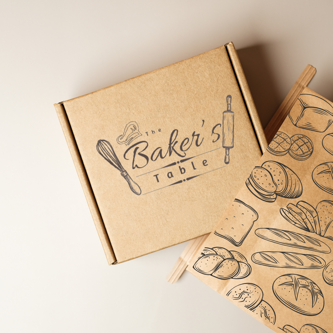 A brown cardboard box labeled 'The Baker's Table' with a whisk and rolling pin illustration. Next to it is a paper bag with line drawings of baked goods and bread.