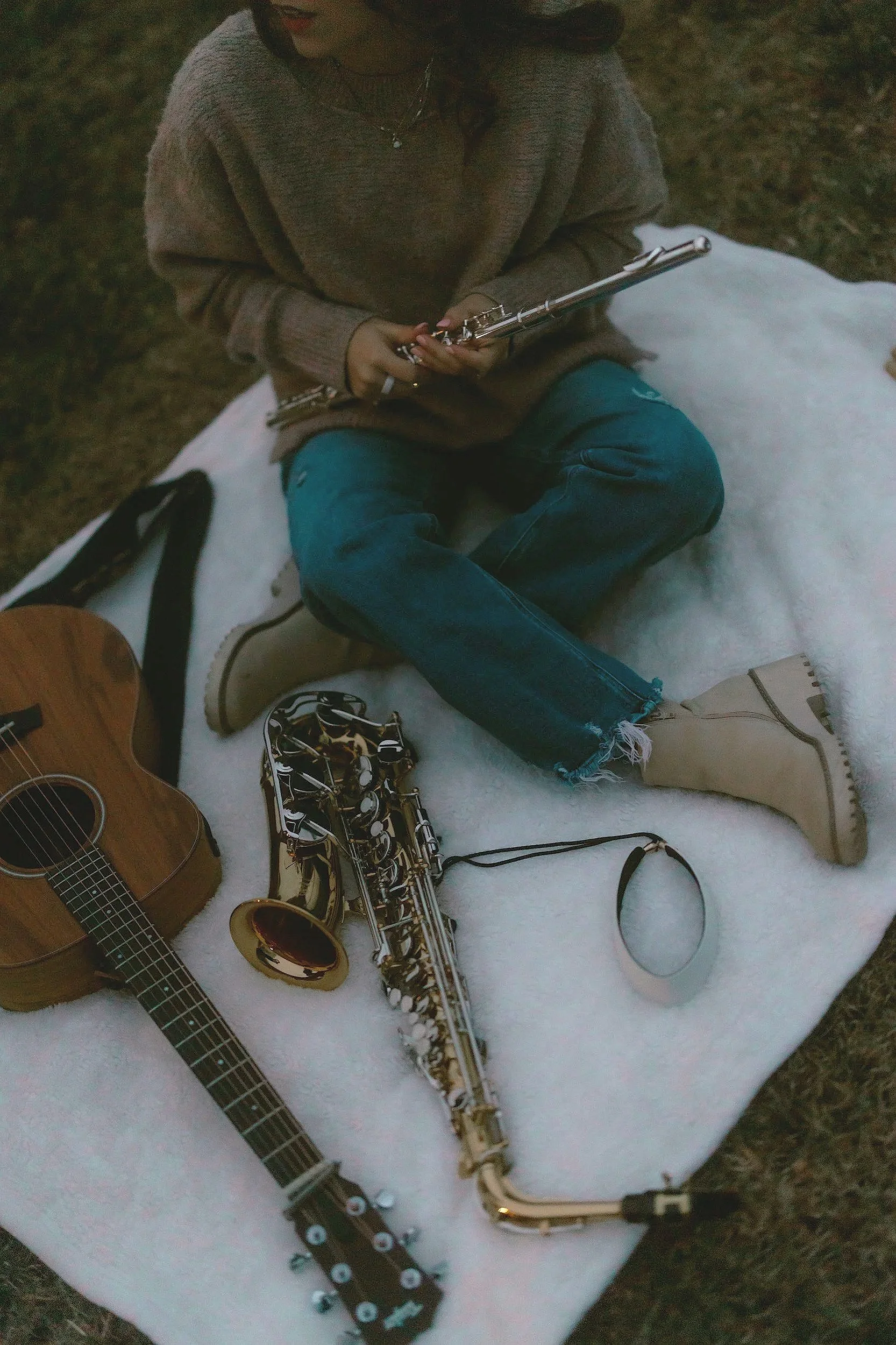Person sitting on a white blanket outdoors, holding a flute. There is an acoustic guitar, a gold saxophone, and a wristband on the blanket.