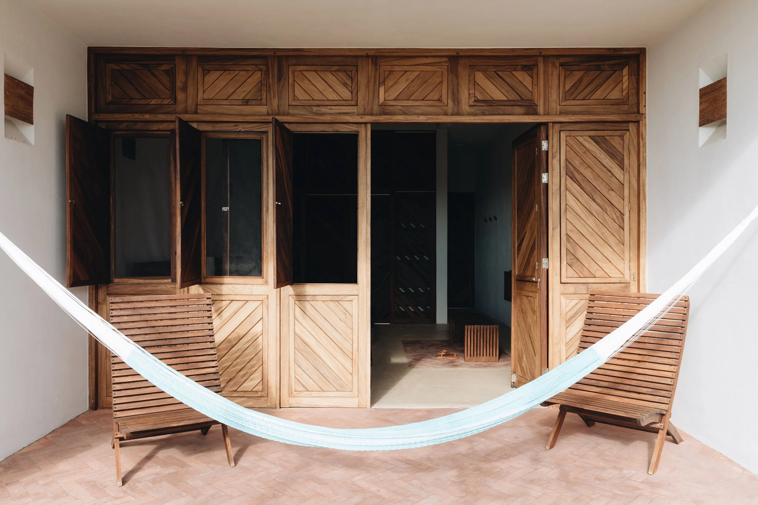 Dos sillones de madera con una hamaca entre ellos en una terraza, con un fondo de armario o pared de madera y paredes blancas.