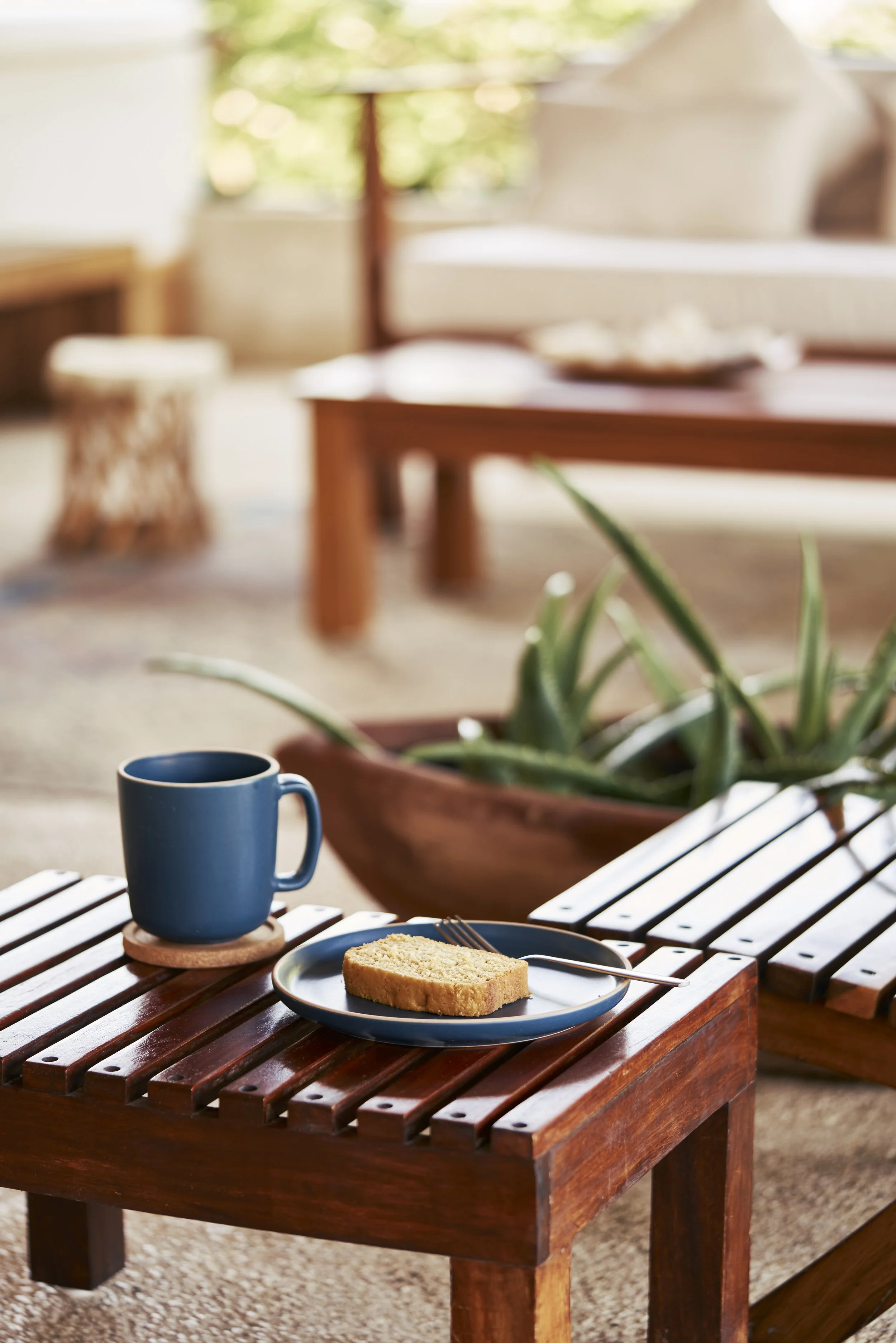 Una taza azul, un plato con un pedazo de pastel y un tenedor en una mesa de madera en un ambiente interior con plantas y mobiliario de madera.