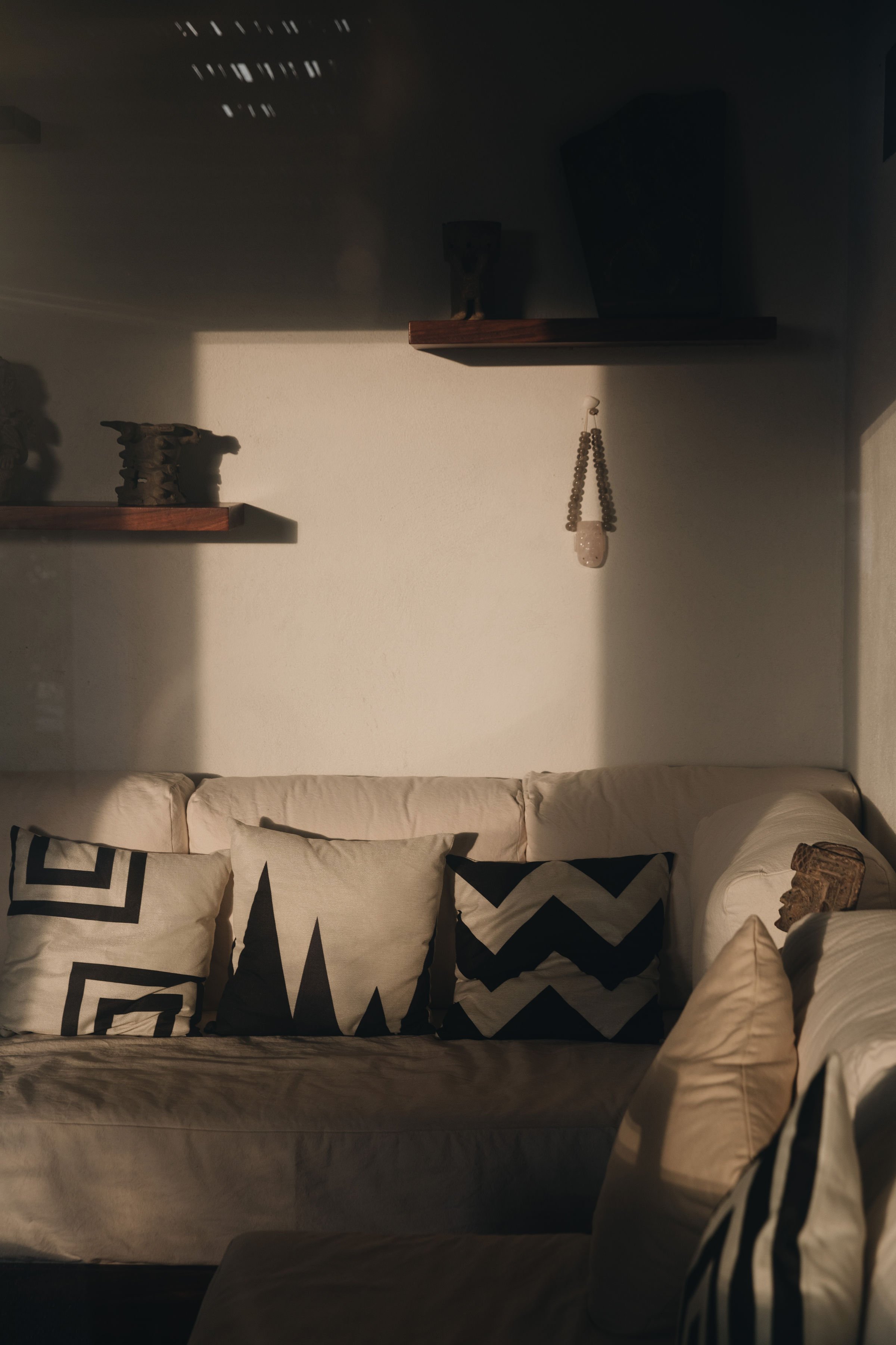 Sala de estar con un sillón beige y varias almohadas con patrones en blanco y negro, y estanterías con objetos decorativos en la pared, iluminada por luz natural.
