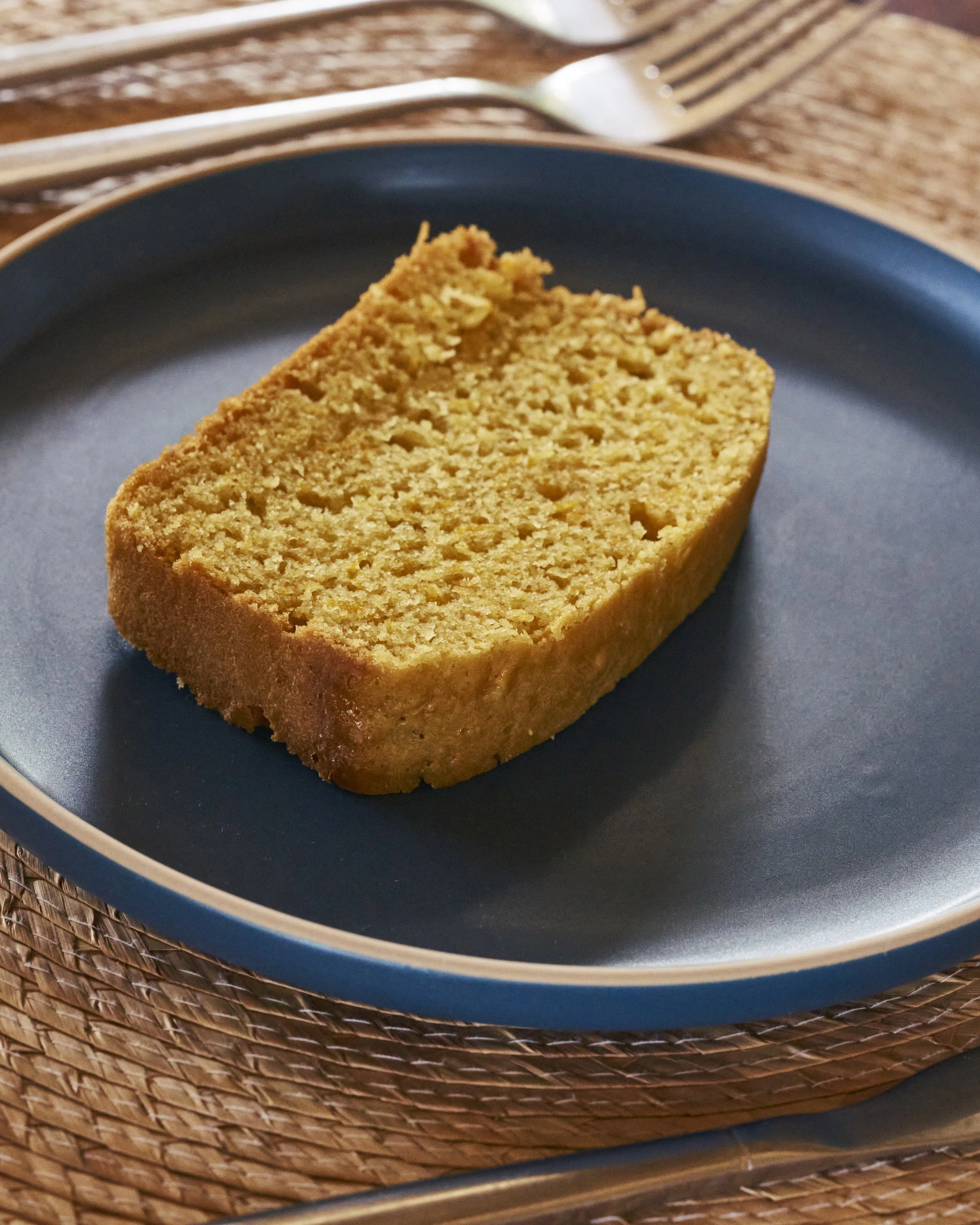 Un pedazo de pastel de vainilla en un plato azul oscuro sobre una mesa de madera, con tenedor y cuchillo al fondo.