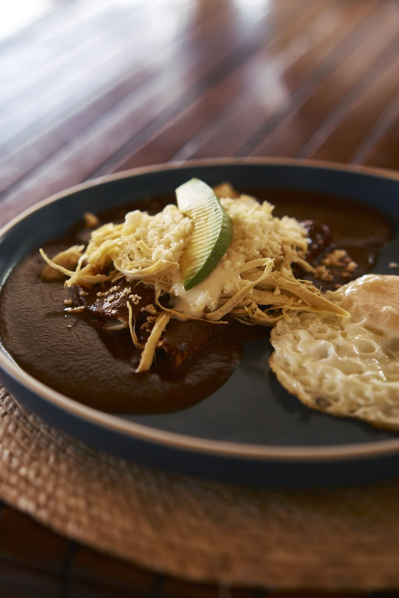 Plato de comida mexicana con carne en salsa, frijoles, arroz, queso, rodaja de limón y huevo estrellado en plato negro sobre mesa de madera.