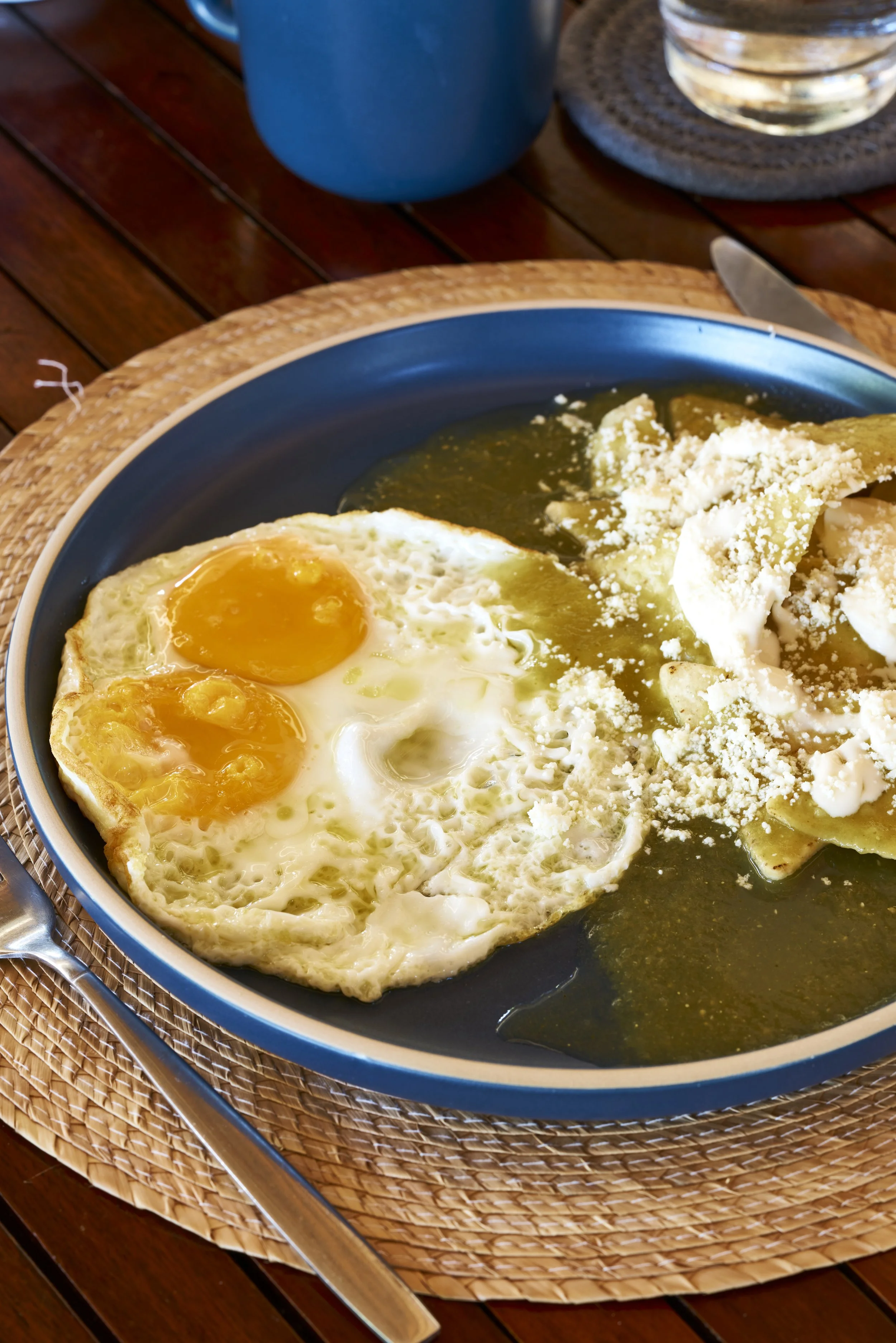 Plato de huevos estrellados, salsa, queso, y tortillas en un plato azul sobre una mesa de madera.