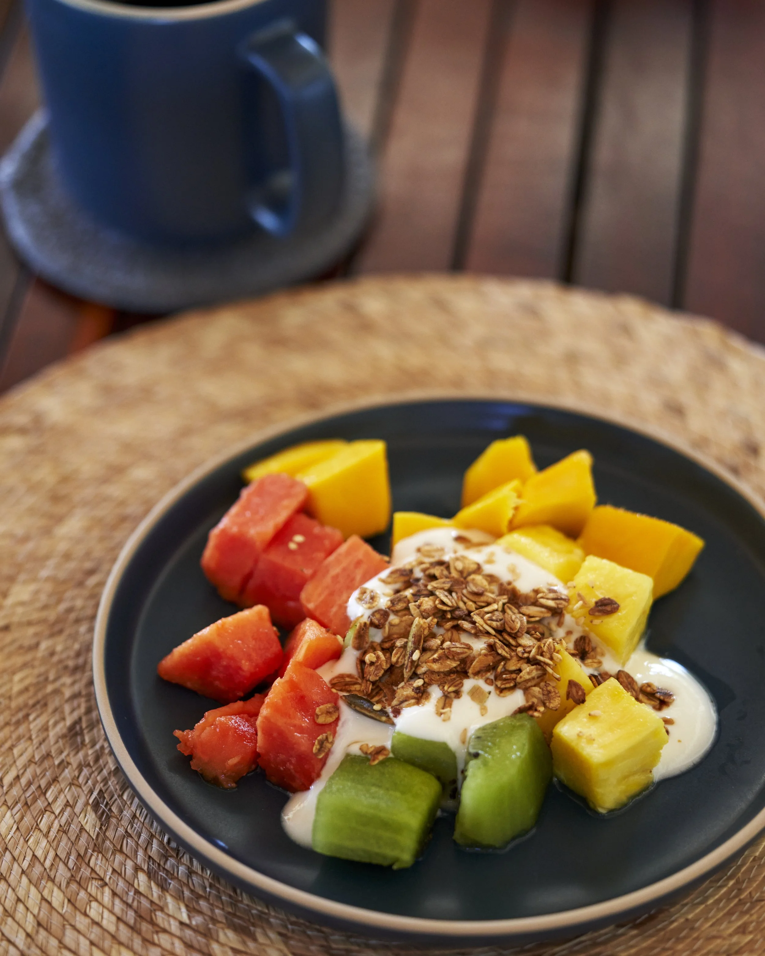 En un plato negro hay rodajas de melón, sandía, piña y papaya con yogur y avena; al fondo, en una mesa de madera, una taza negra en un plato de ratán.
