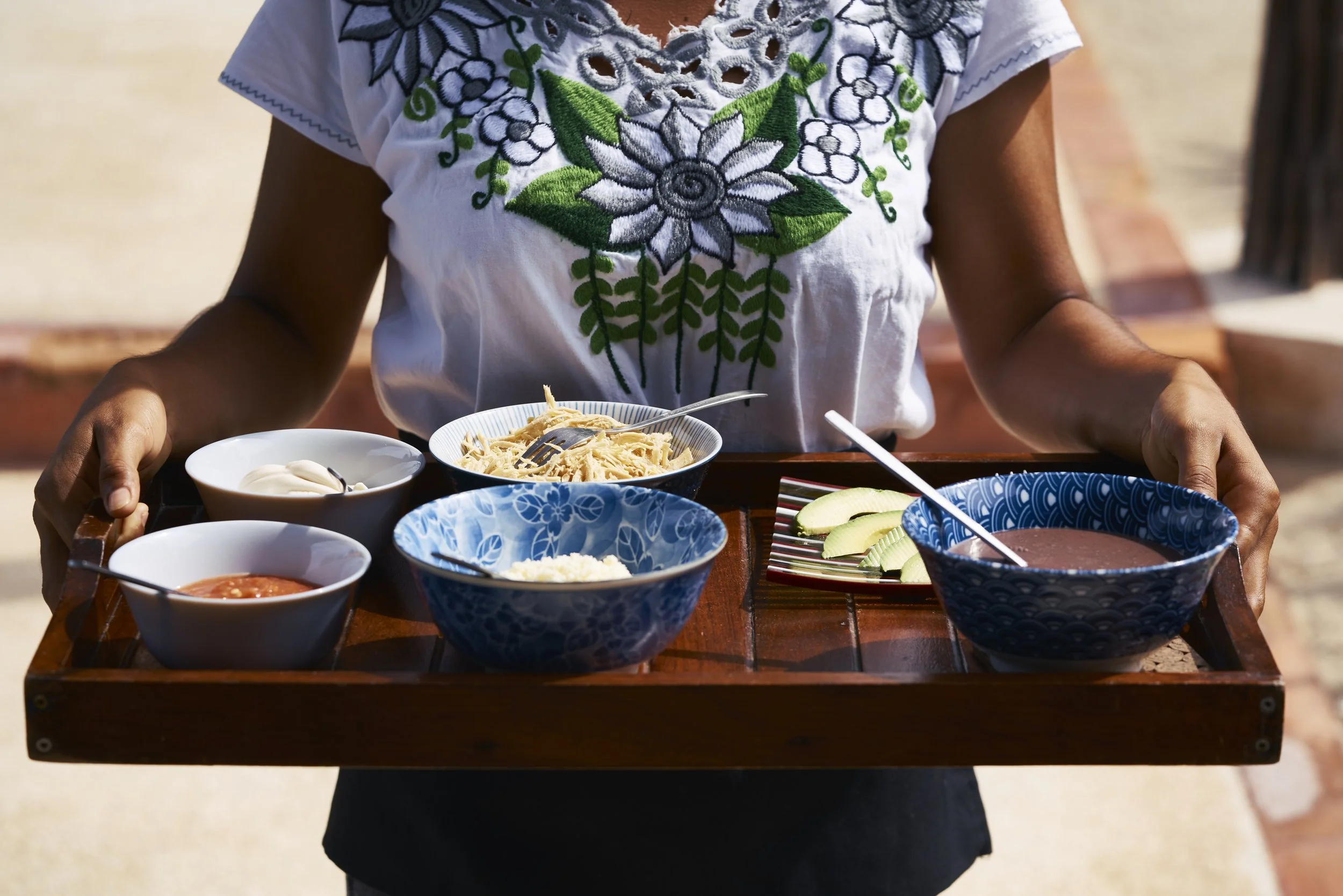 Persona sosteniendo una bandeja con varios platos de comida mexicana, incluyendo salsas, pasta, frutas y crema, en un ambiente al aire libre.