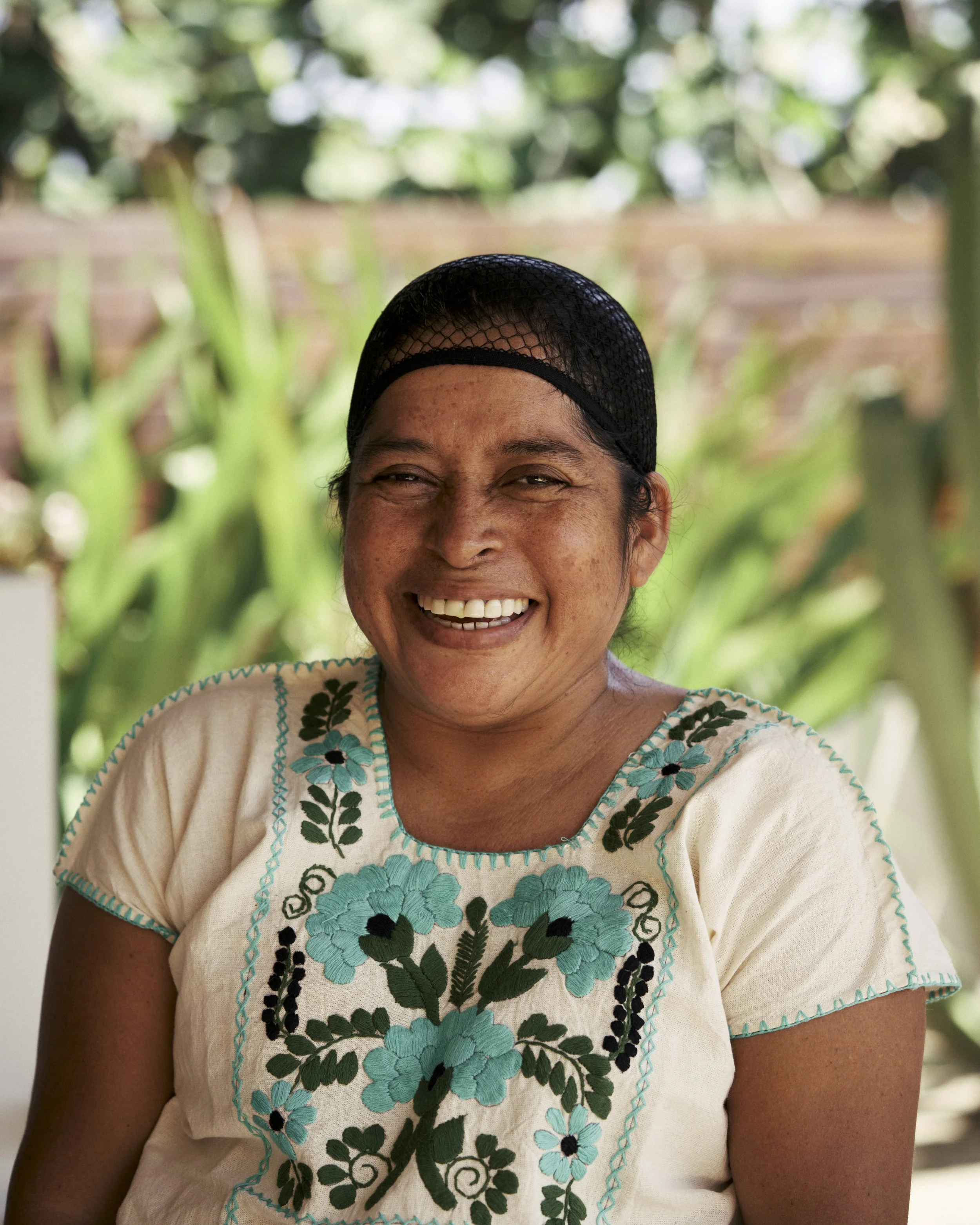Mujer sonriente con sombrero de red, camiseta beige con bordados florales en tonos azules y verdes, fondo de plantas verdes.