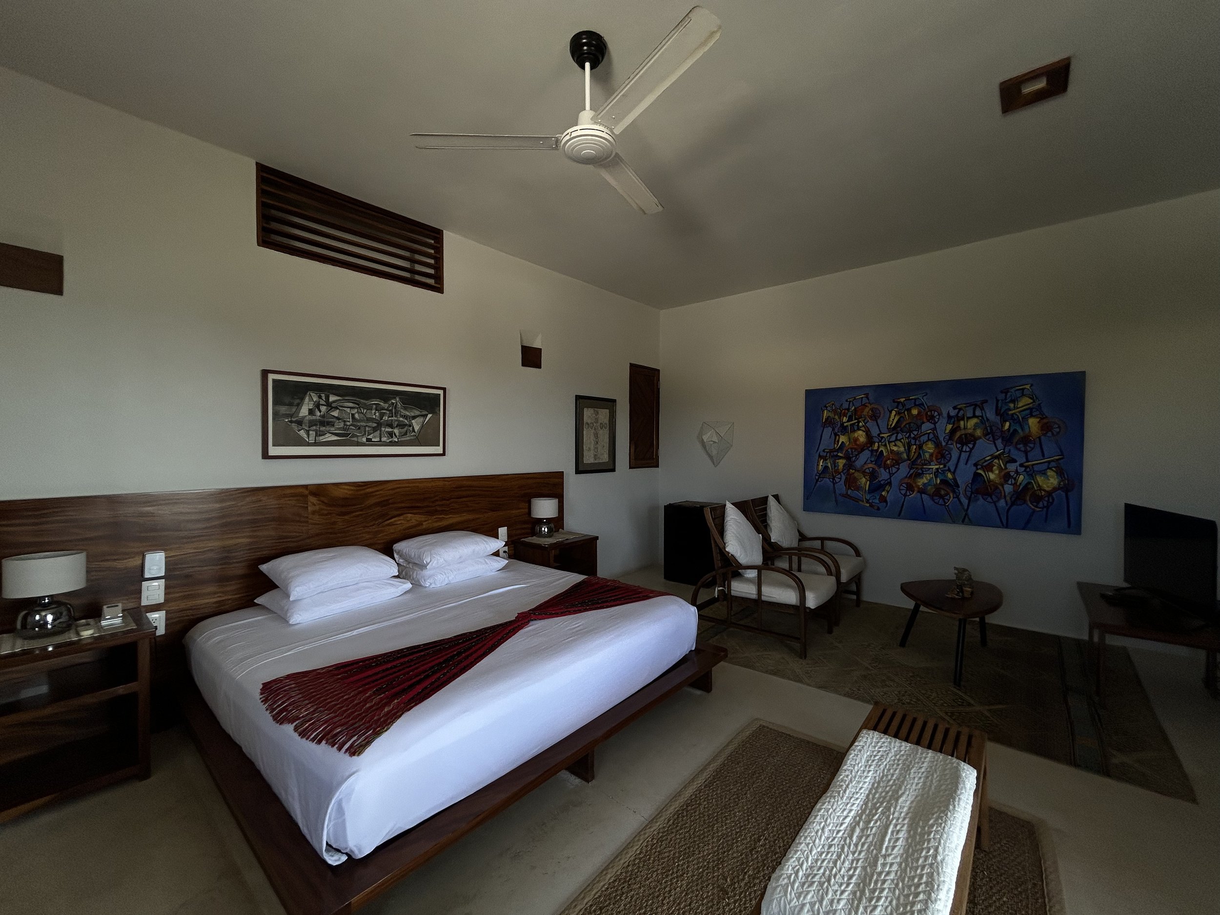 Habitación de hotel con cama grande, mesa de noche con lámpara, sillones, mesa pequeña, televisión y arte en las paredes.