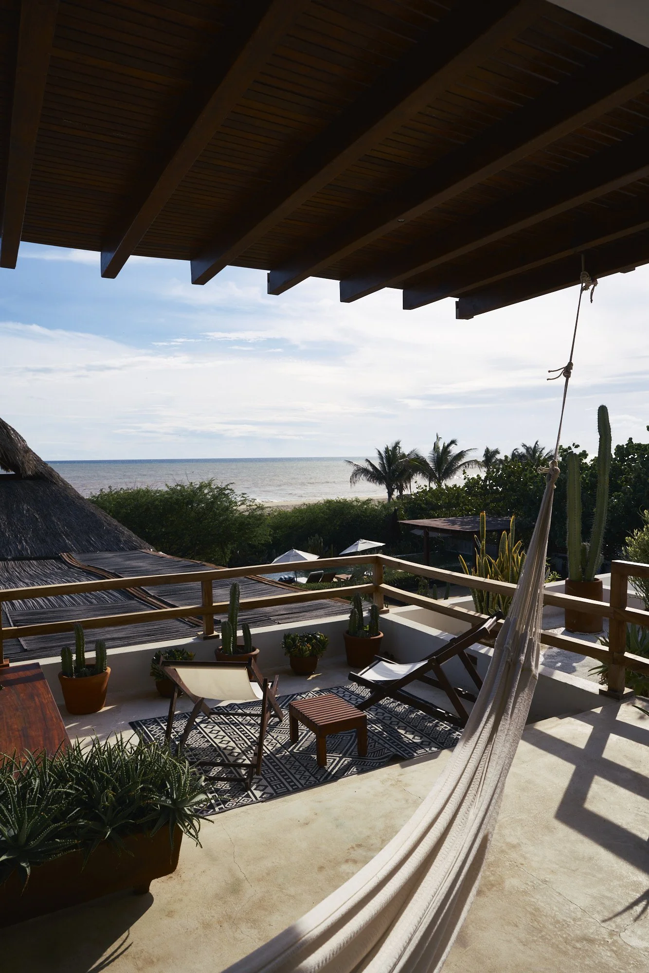 Vista de una terraza con hamaca, sillas, plantas en macetas y vista al mar con palmeras en el fondo, en un día soleado y despejado.