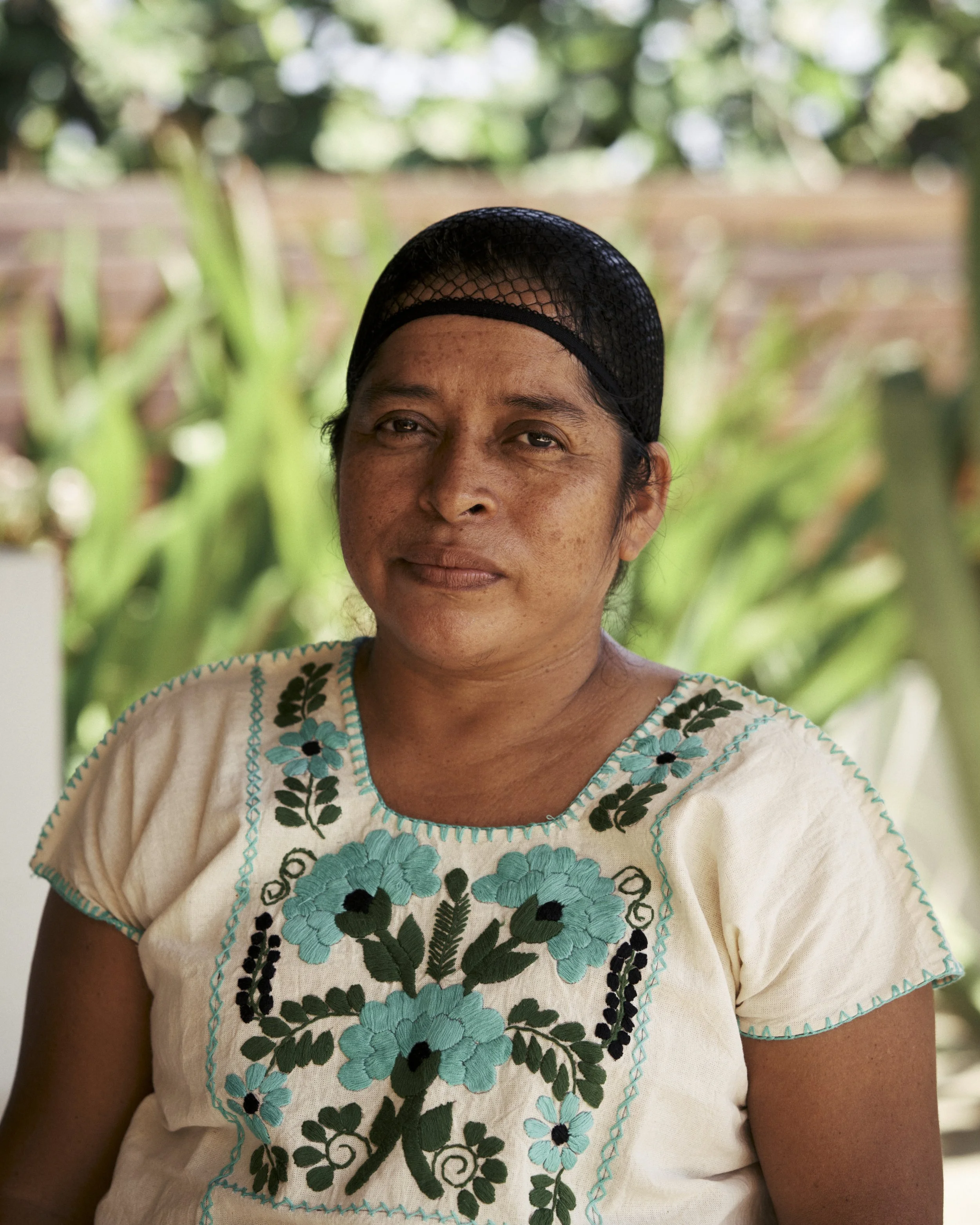Retrato de una mujer con gorra de red y blusa bordada con flores azul y verde, fondo con plantas verdes.