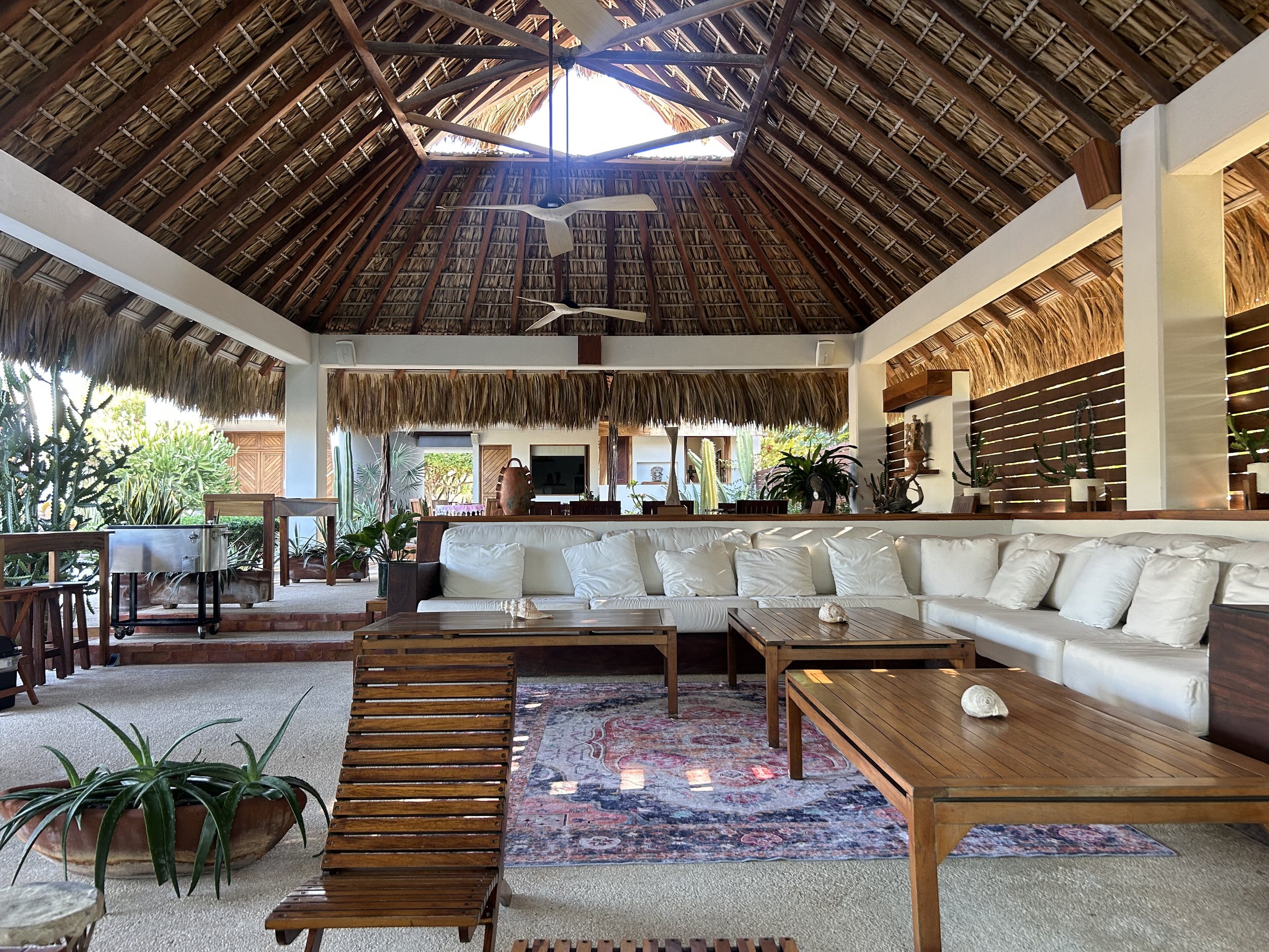 Interior de una sala con techo de palma y ventiladores, con muebles de madera y sofá blanco, decorada con plantas y una alfombra colorida.