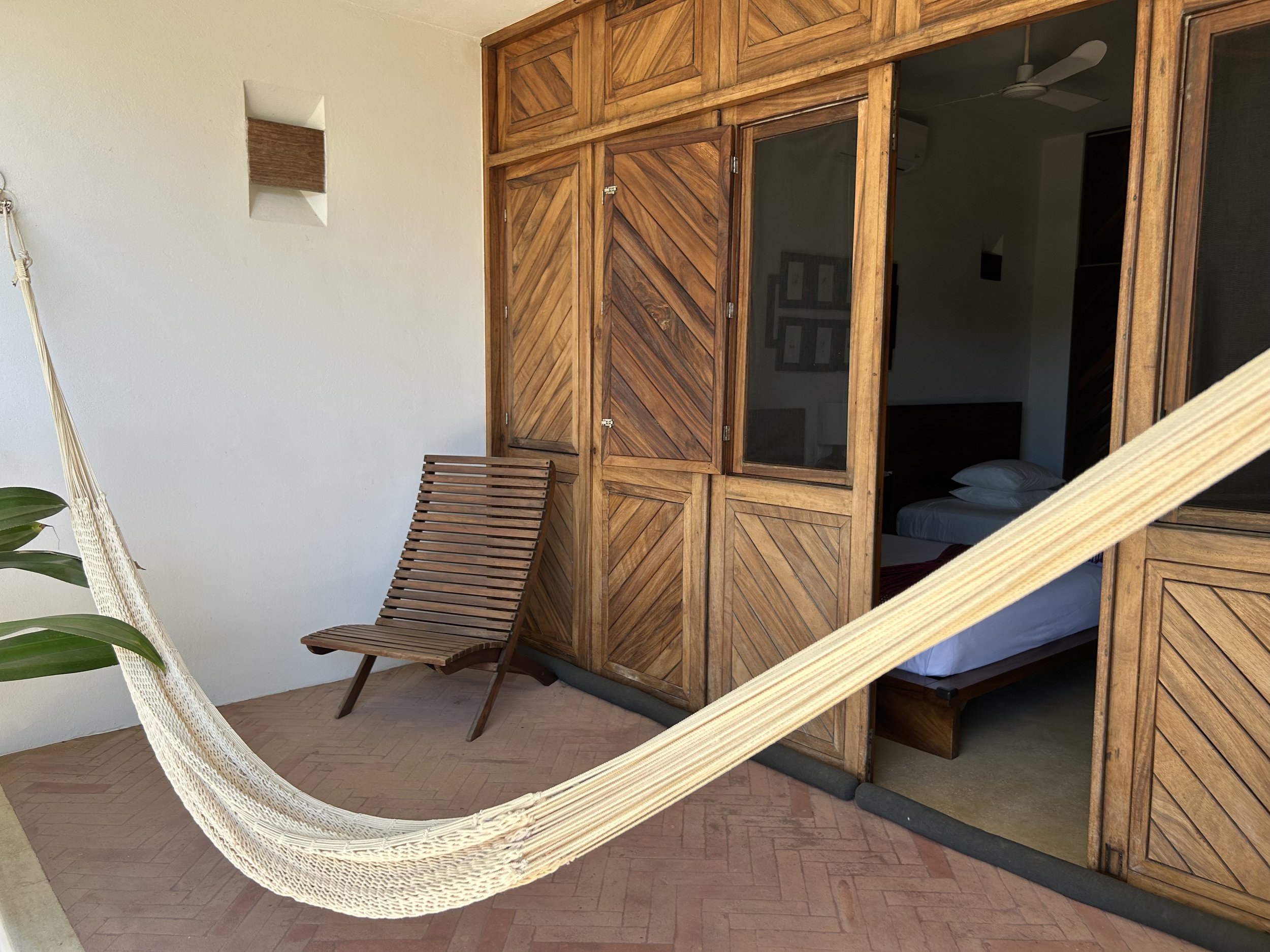 Balcón con hamaca de fibra natural, silla de madera y puertas de madera hacia una habitación con cama y cuadros en la pared.
