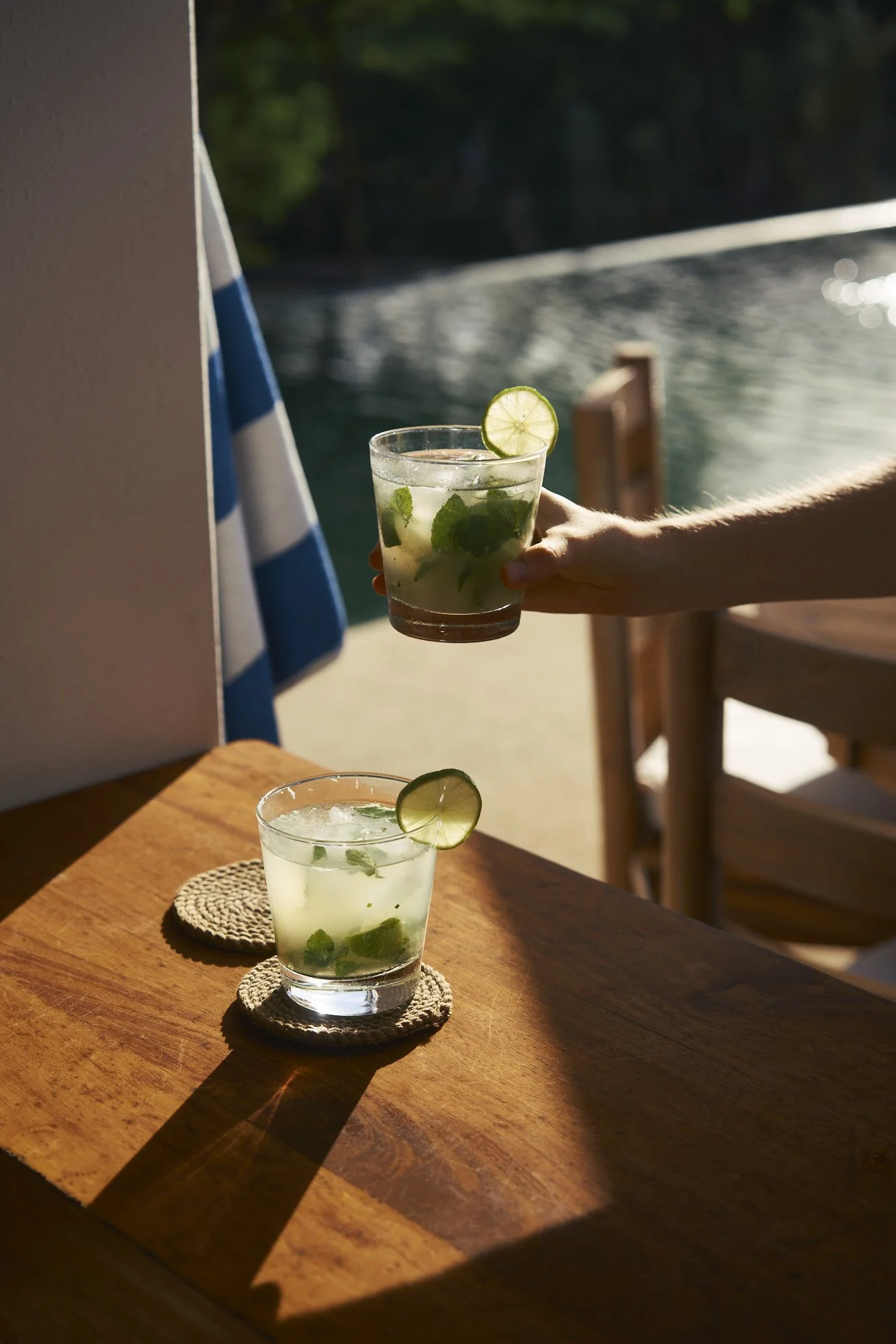 Dos vasos con agua con hielo, rodajas de limón y hierbas, uno sobre una mesa de madera y otro siendo entregado por una mano, con fondo de agua y naturaleza.