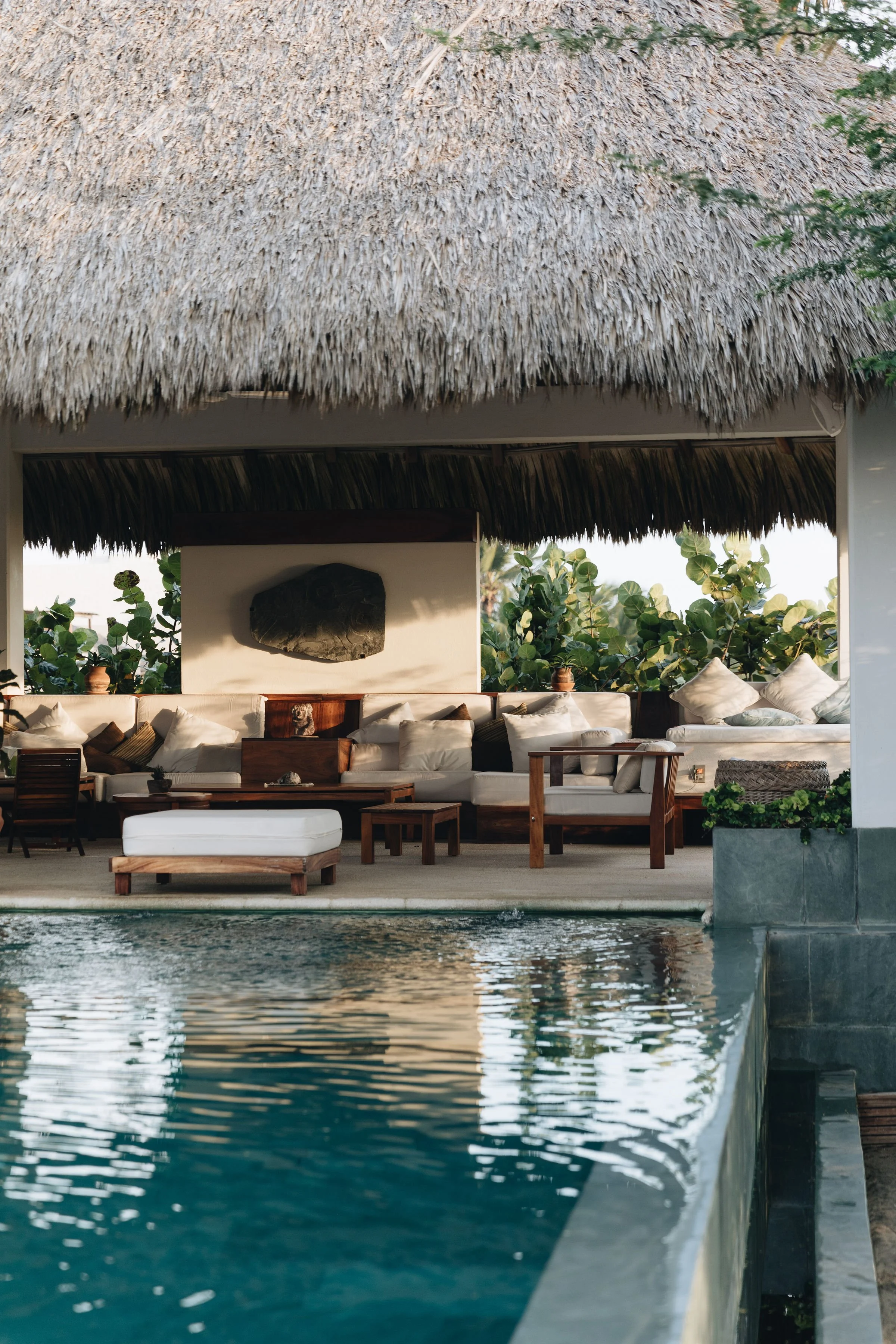 Área de descanso con sillones y mesas de madera junto a una piscina, con techo de palapa y plantas alrededor.