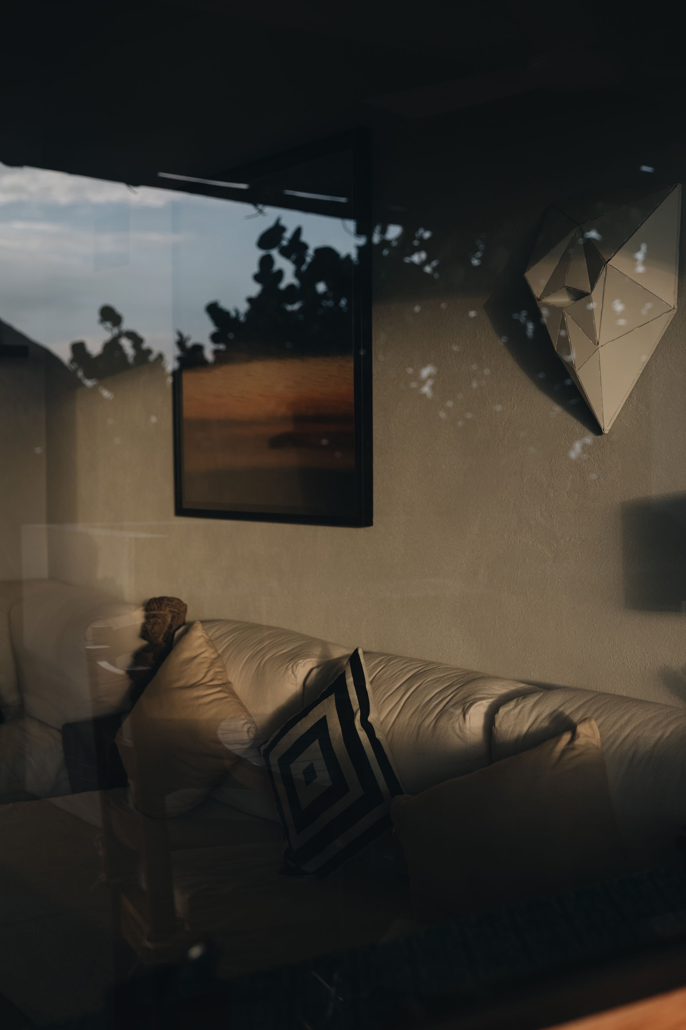 Vista de un sofá beige con cojines, una pared con un cuadro y una escultura de formas geométricas, y una ventana que refleja árboles y el cielo.