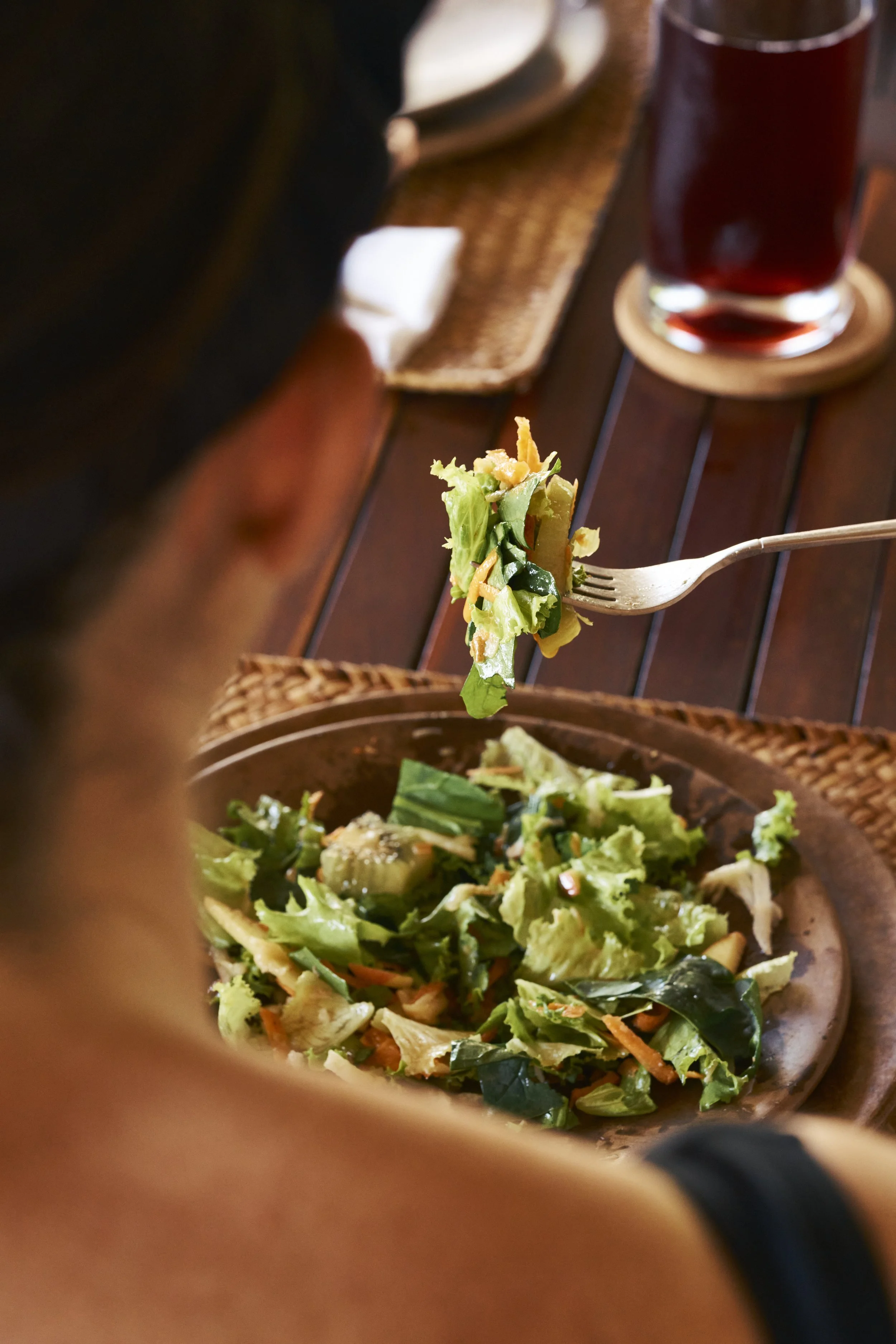 Persona comiendo ensalada en un plato de cerámica en una mesa de madera. En el fondo, una bebida oscura y un lugar de comida.