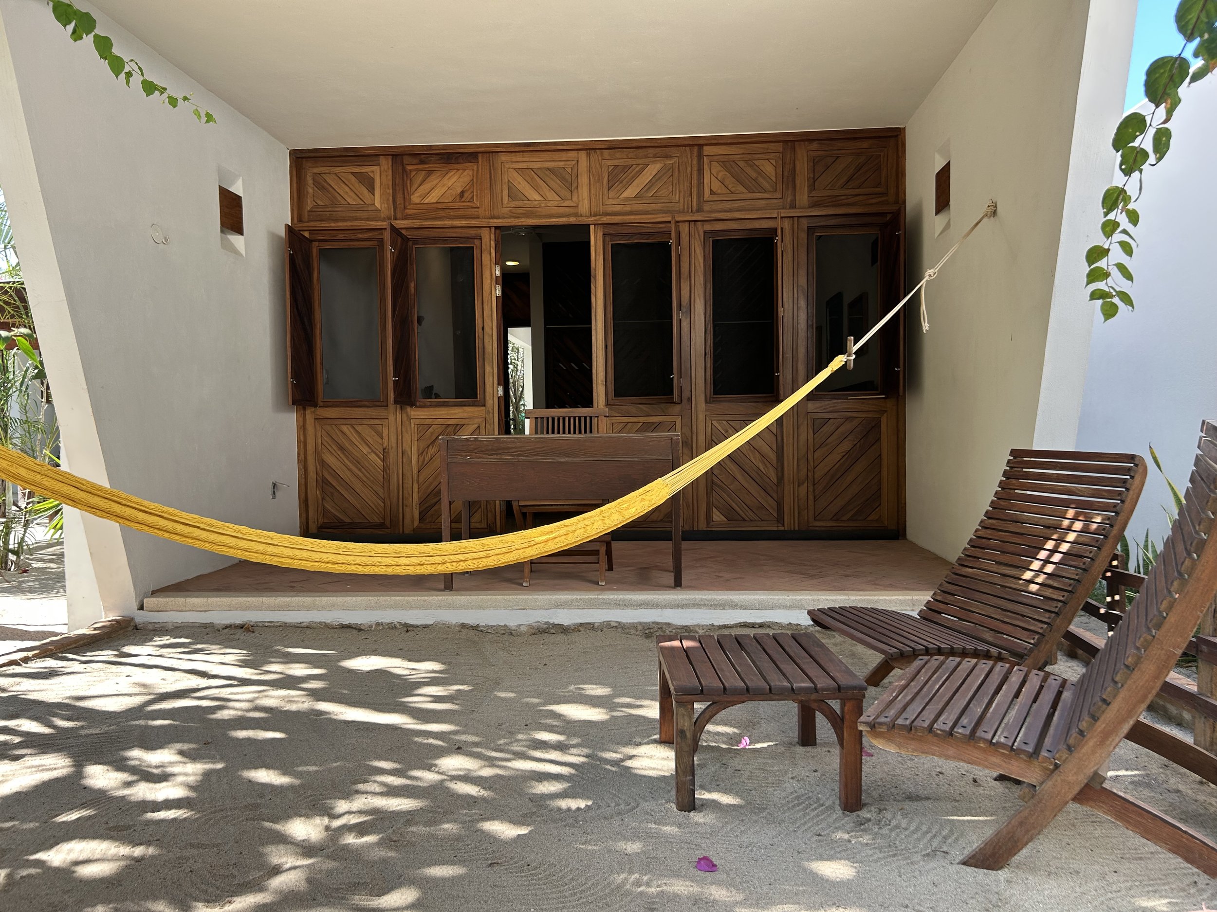 Sombra de árbol en la arena, dos sillones de madera y una hamaca amarilla en un espacio exterior moderno con paredes blancas y una estructura de madera.