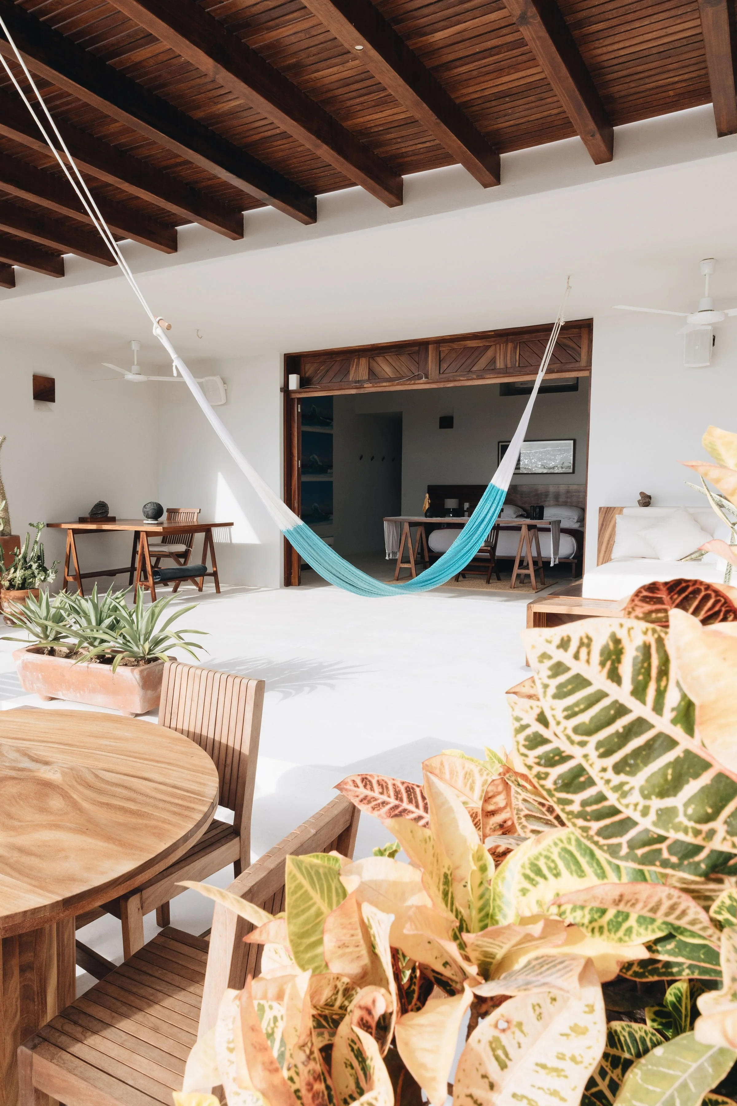 Interior de una sala de estar con muebles de madera, plantas decorativas y una hamaca en el centro, con techo de madera y paredes blancas.