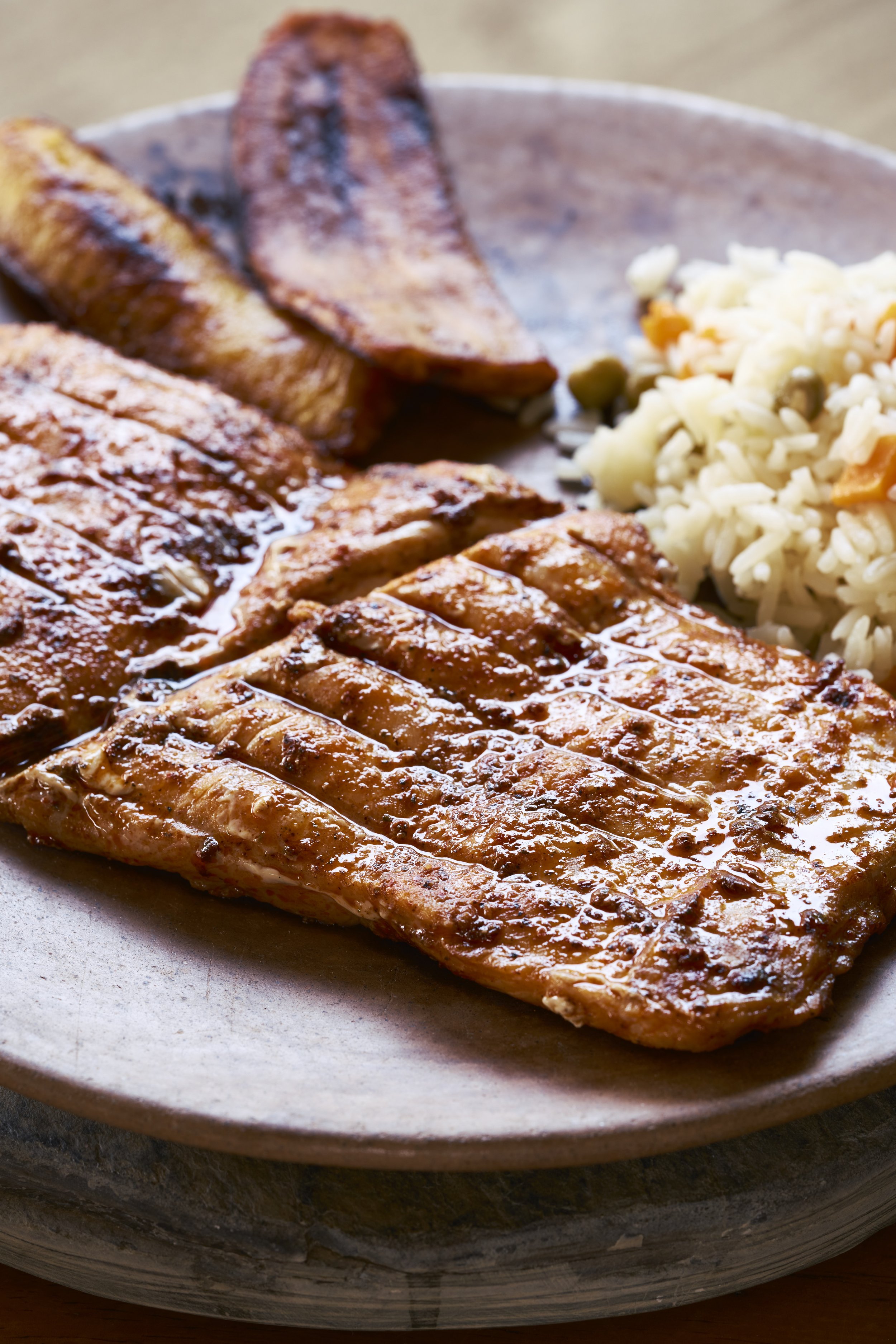 Pechuga de pollo a la parrilla con salsa, plátanos fritos y ensalada de arroz con verduras en plato de cerámica.