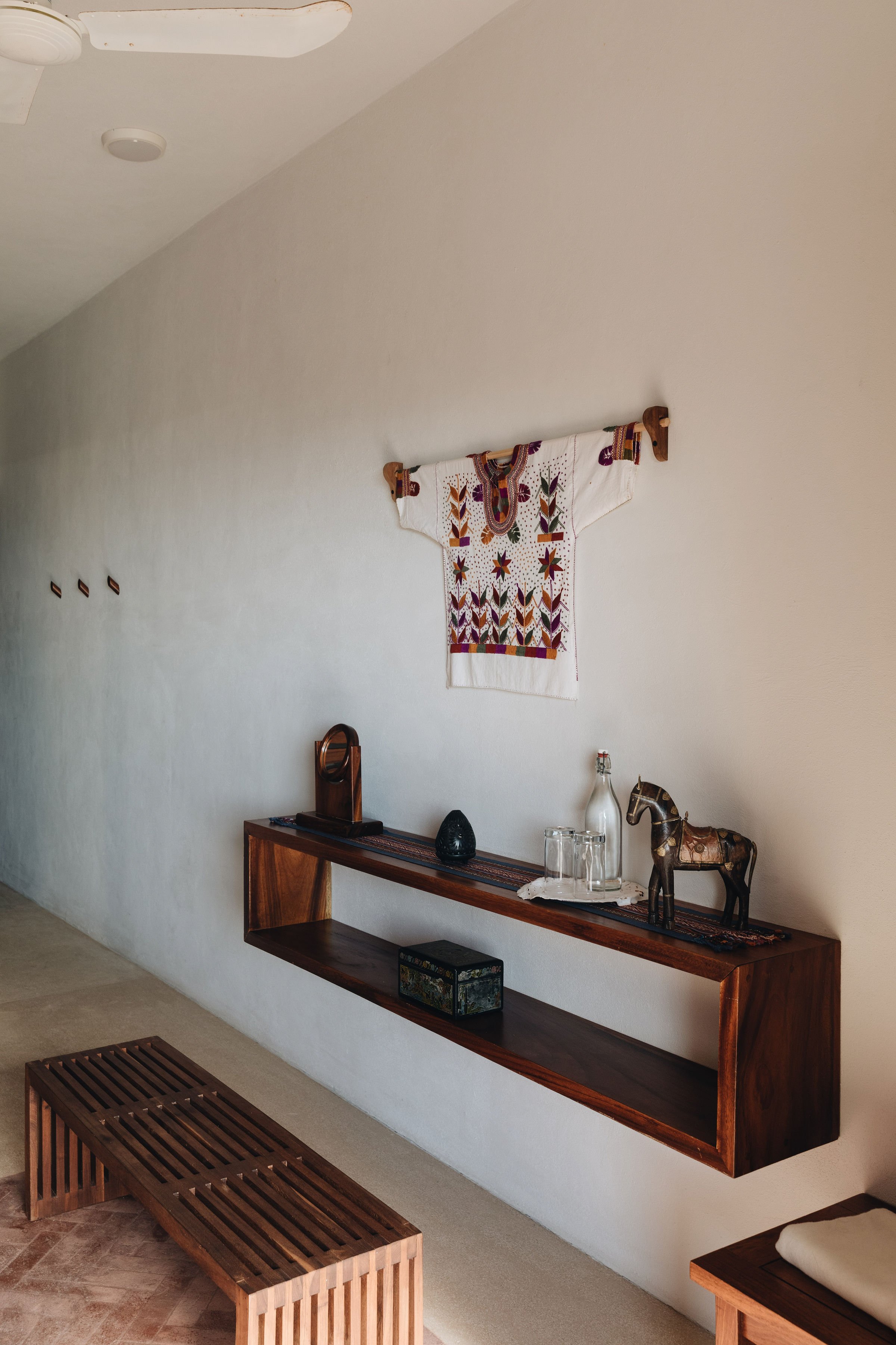 Decoración de estilo étnico en una pared blanca con un pulso de madera flotante. En la pared, un tejido bordado colorido cuelga en la parte superior derecha. Sobre el pulso, hay varias decoraciones: una botella de vidrio, un recipiente de cerámica, u