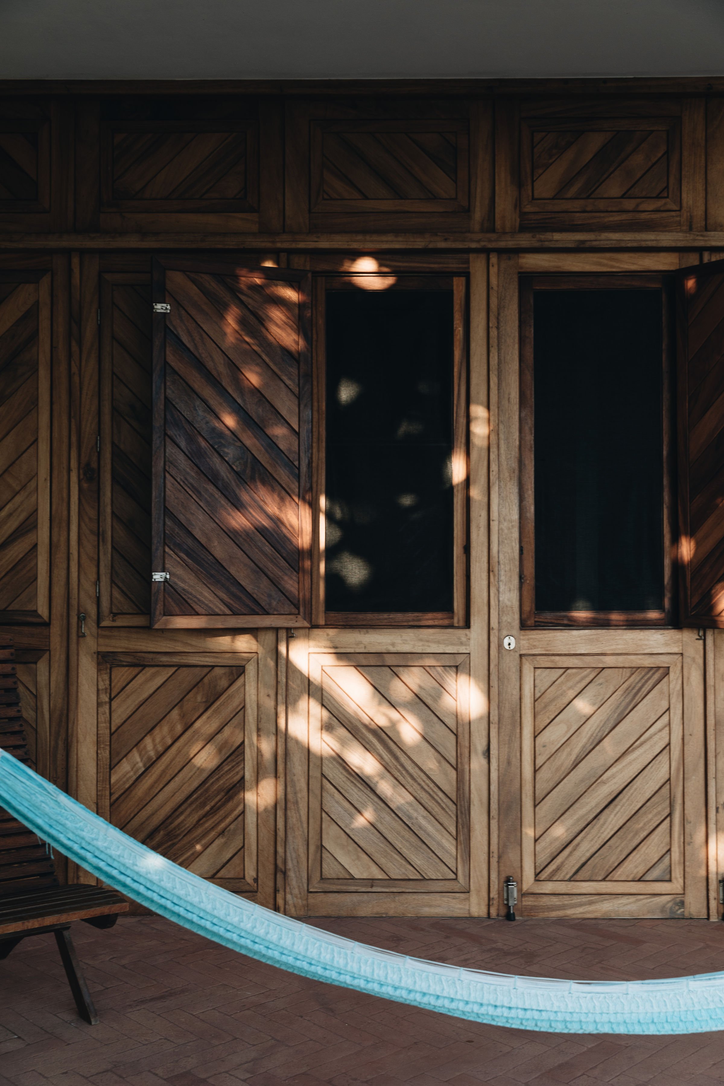 Puerta de madera con persianas en un fondo oscuro, con luz y sombra de hojas en la superficie, y una hamaca azul en primer plano.