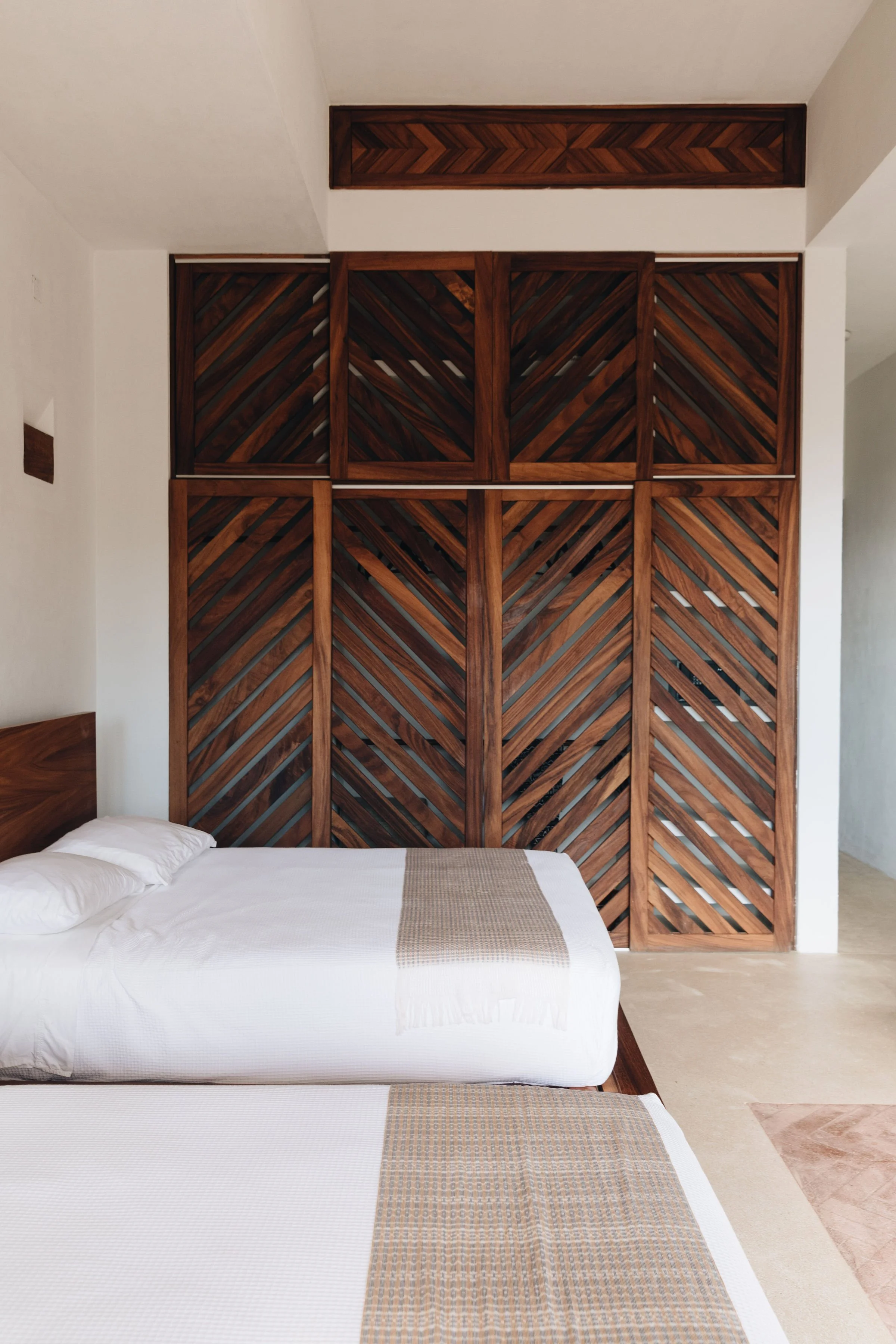 Habitación con paredes blancas, cabecera de madera, camas con sábanas blancas y mantas beige, pared decorativa de madera con patrón de vetas diagonales, suelo de piedra clara.