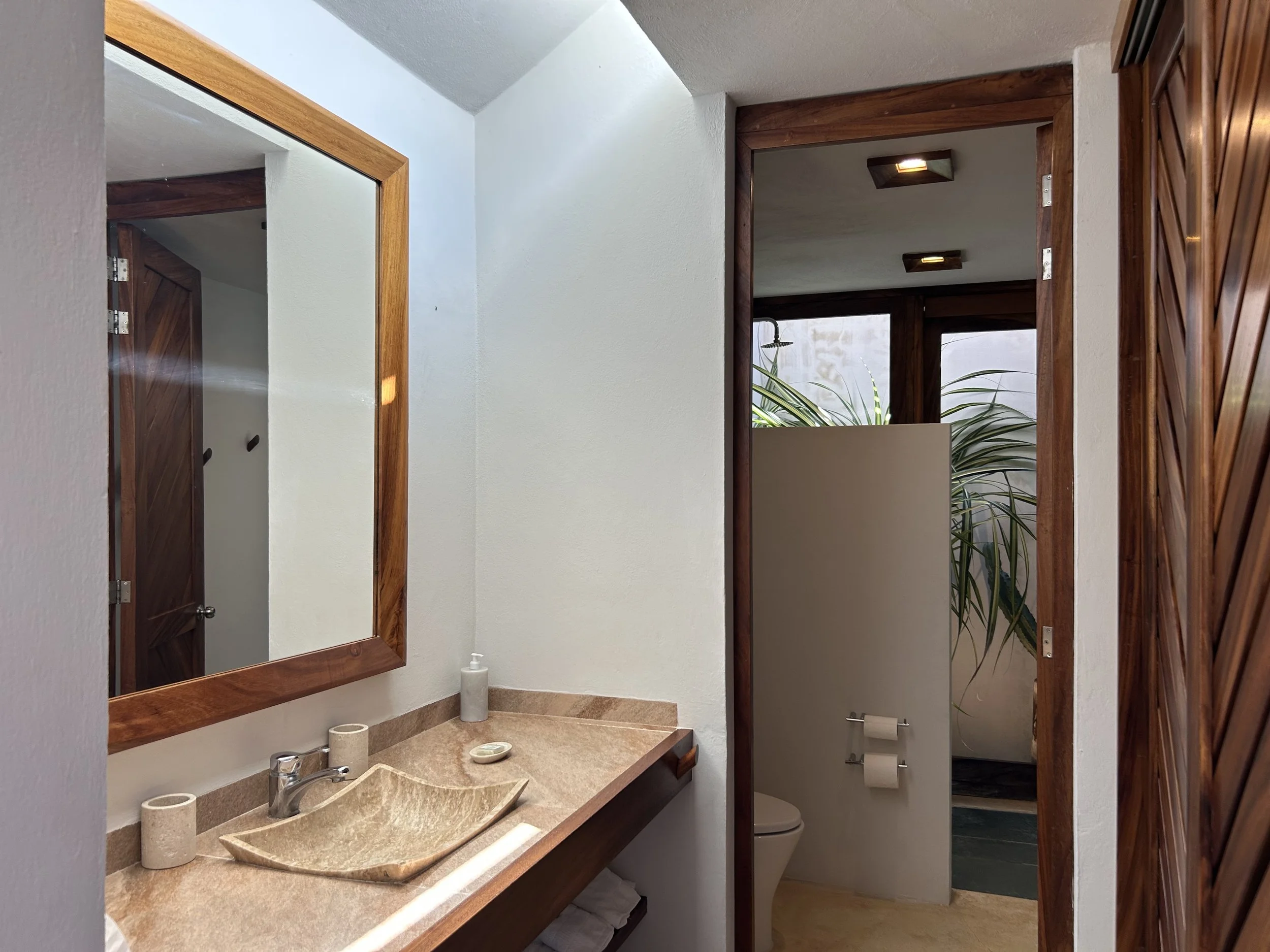 Vista de un baño con lavabo de piedra, espejo, y puerta que lleva a un área con plantas y inodoro, decorado con madera y paredes blancas.