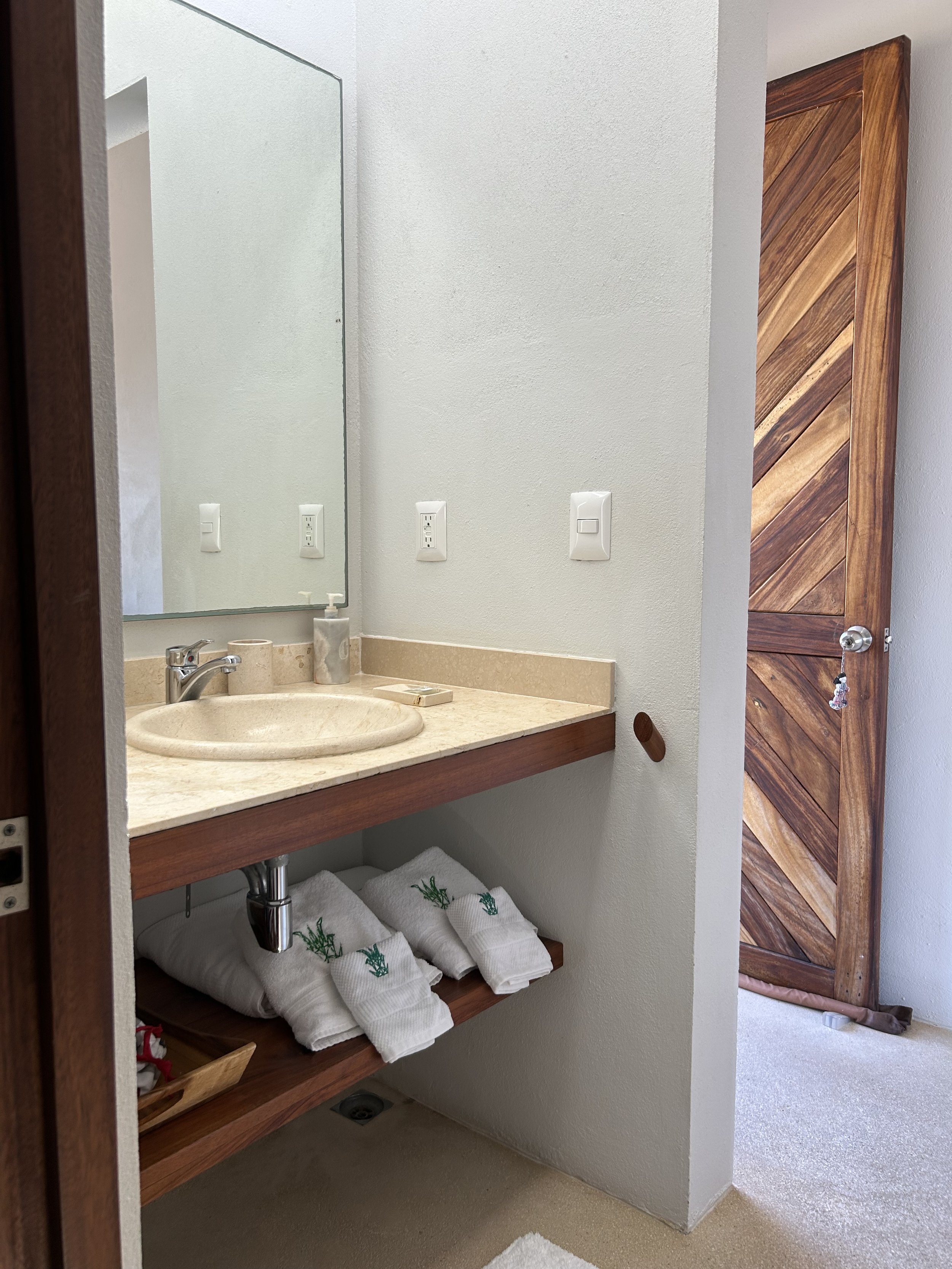 Lavabo con encimera de piedra, espejo grande, jabonera y toallas blancas con símbolos de cannabis en un pasillo de baño con puerta de madera a la derecha.