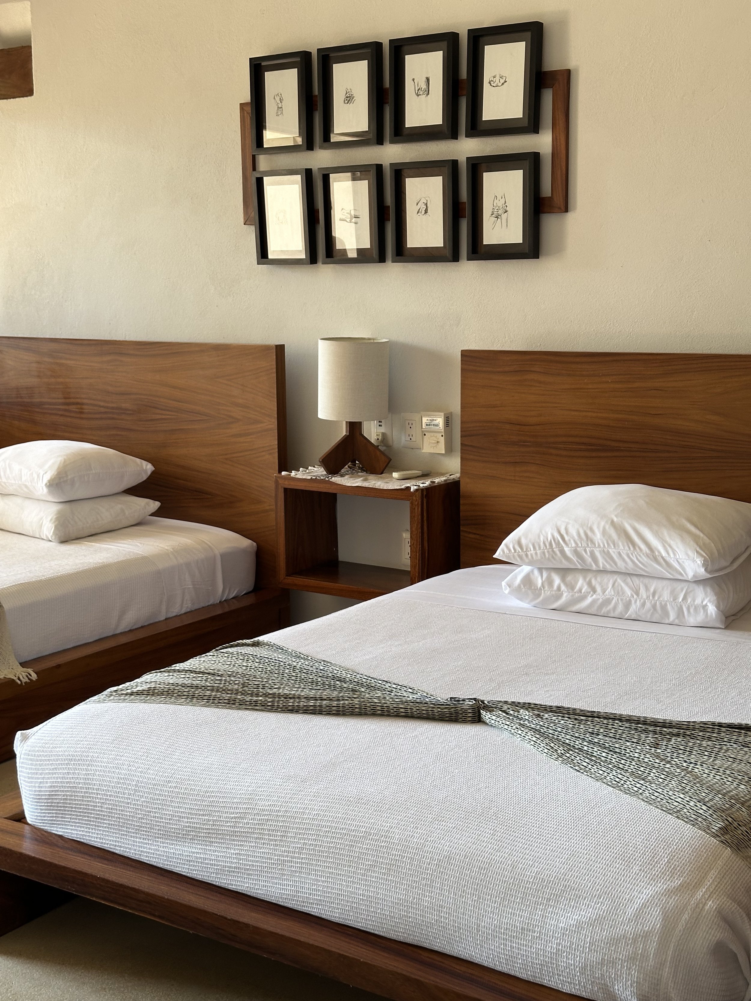 Habitación con dos camas de madera, una con almohadas blancas y ropa de cama blanca, dentro de una habitación con una pared beige, un cabecero de madera, una lámpara de noche y un cuadro con varias imágenes enmarcadas en negro y madera.