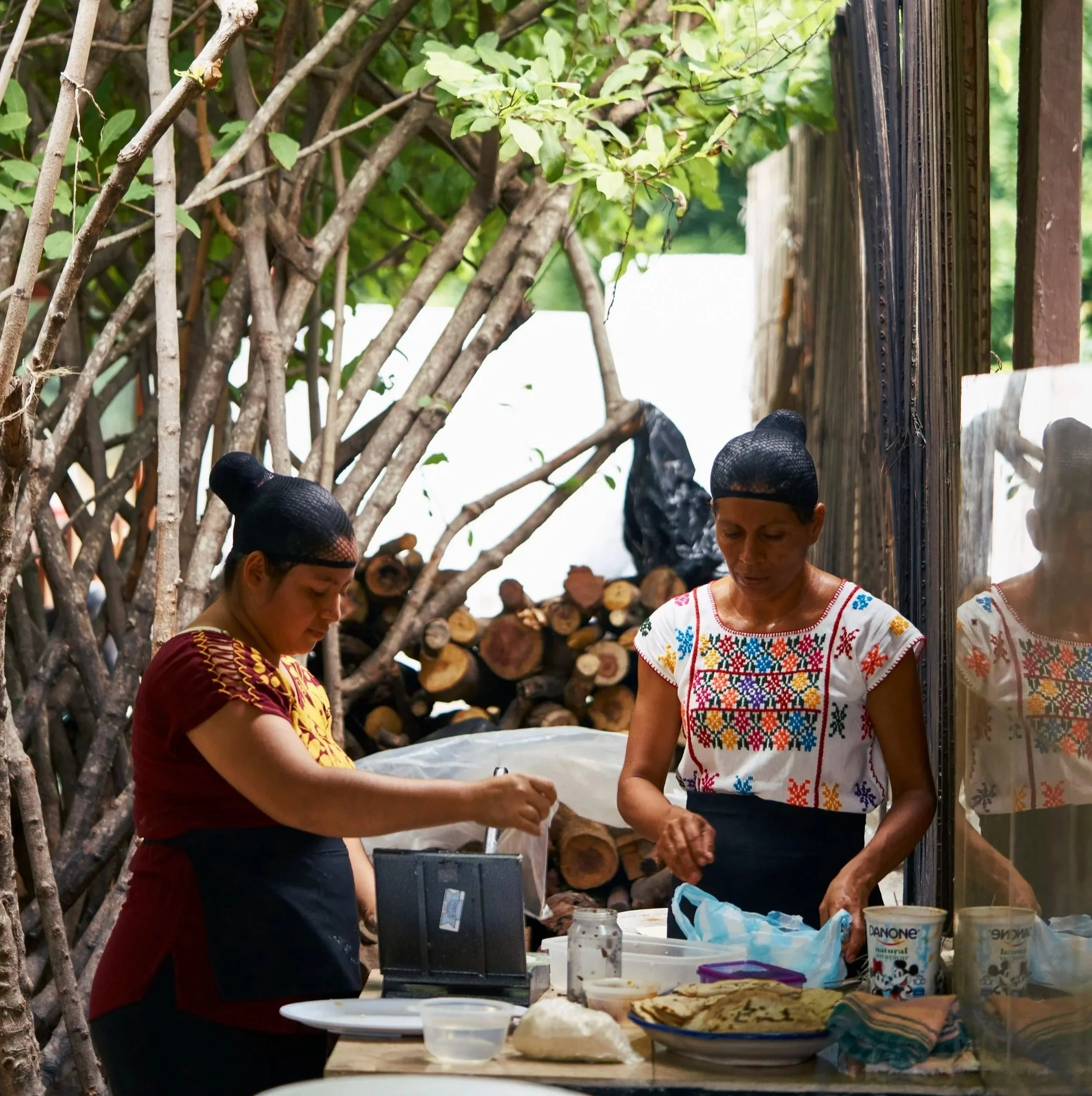 Dos mujeres indígenas preparando comida en un puesto al aire libre, rodeadas de madera y vegetación.