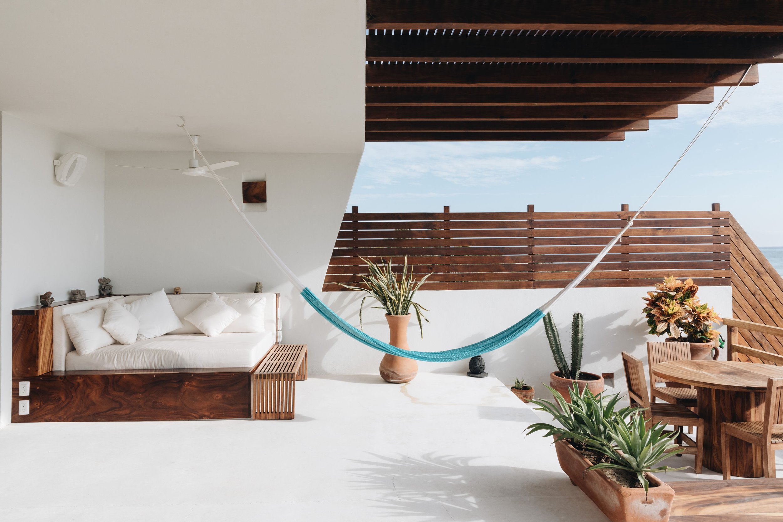 Terraza con hamaca azul, sillón de madera, plantas en macetas y vista al mar.