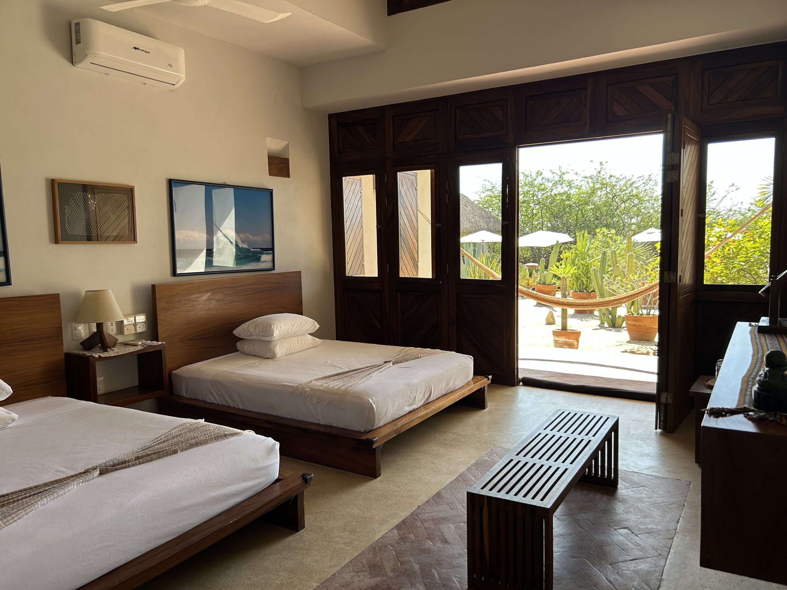 Habitación con dos camas individuales, noche de madera, lámpara, pintura de océano, puerta de madera al balcón con plantas y hamaca afuera.