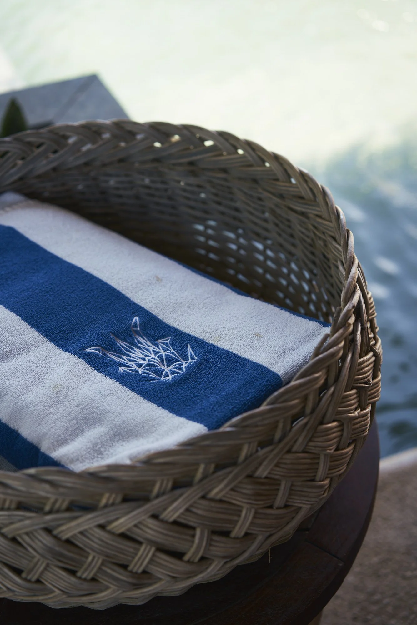 Una toalla blanca con una franja azul, con un logo de un árbol de cristal, dentro de una cesta de mimbre junto a un cuerpo de agua en un entorno tranquilo.
