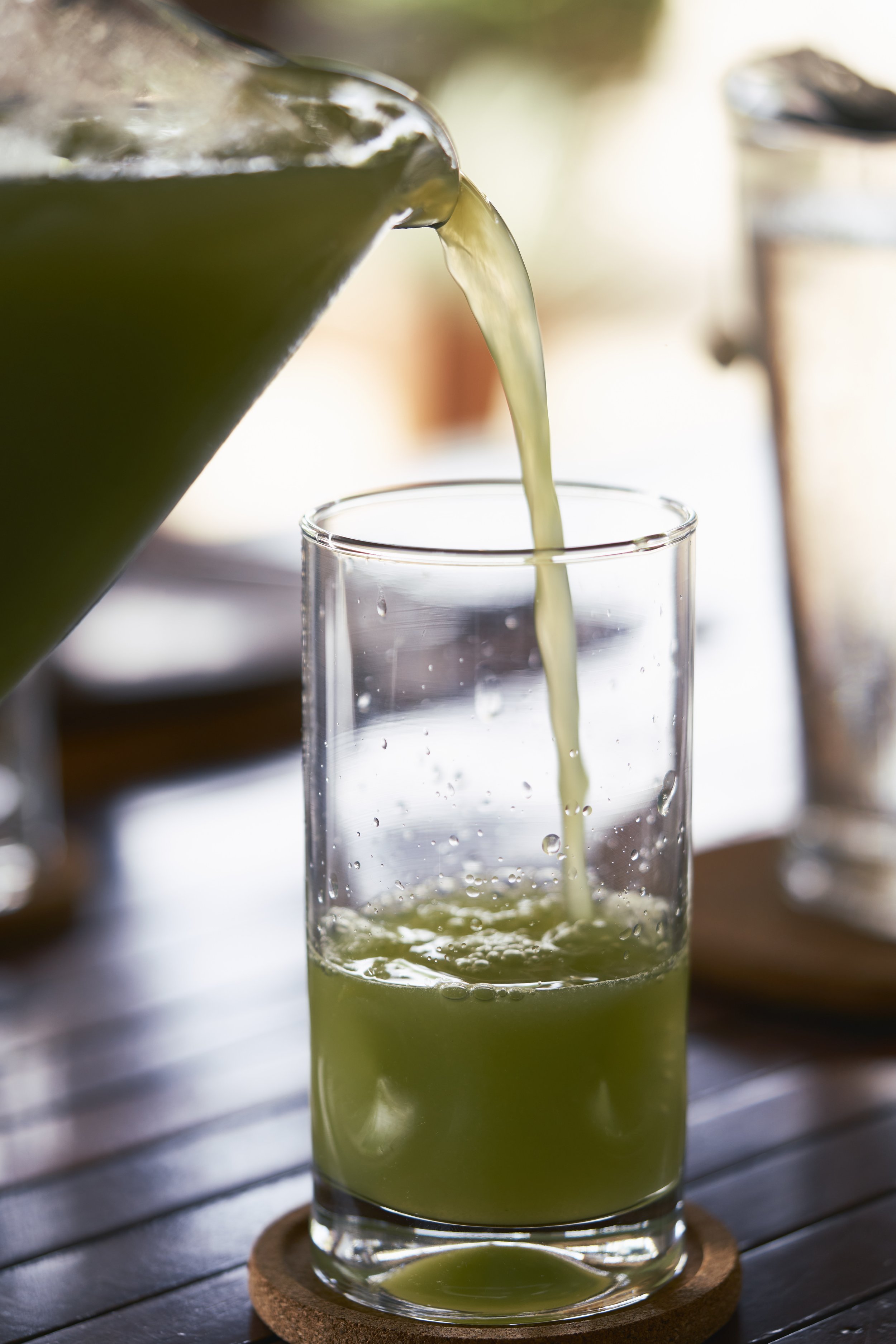 Vaso siendo llenado con jugo verde desde una jarra sobre una mesa de madera.