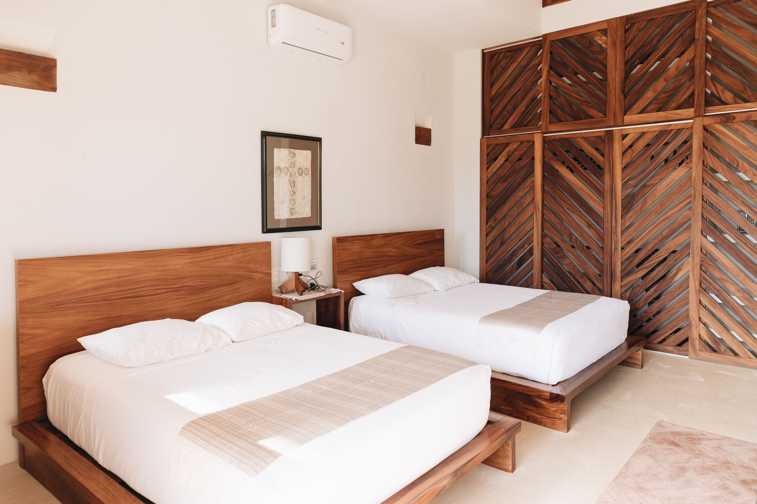 Habitación de hotel con dos camas grandes de madera, una lámpara en una mesa entre las camas, una pared blanca con arte decorativo y una pared con paneles de madera decorativos, aire acondicionado en la parte superior.