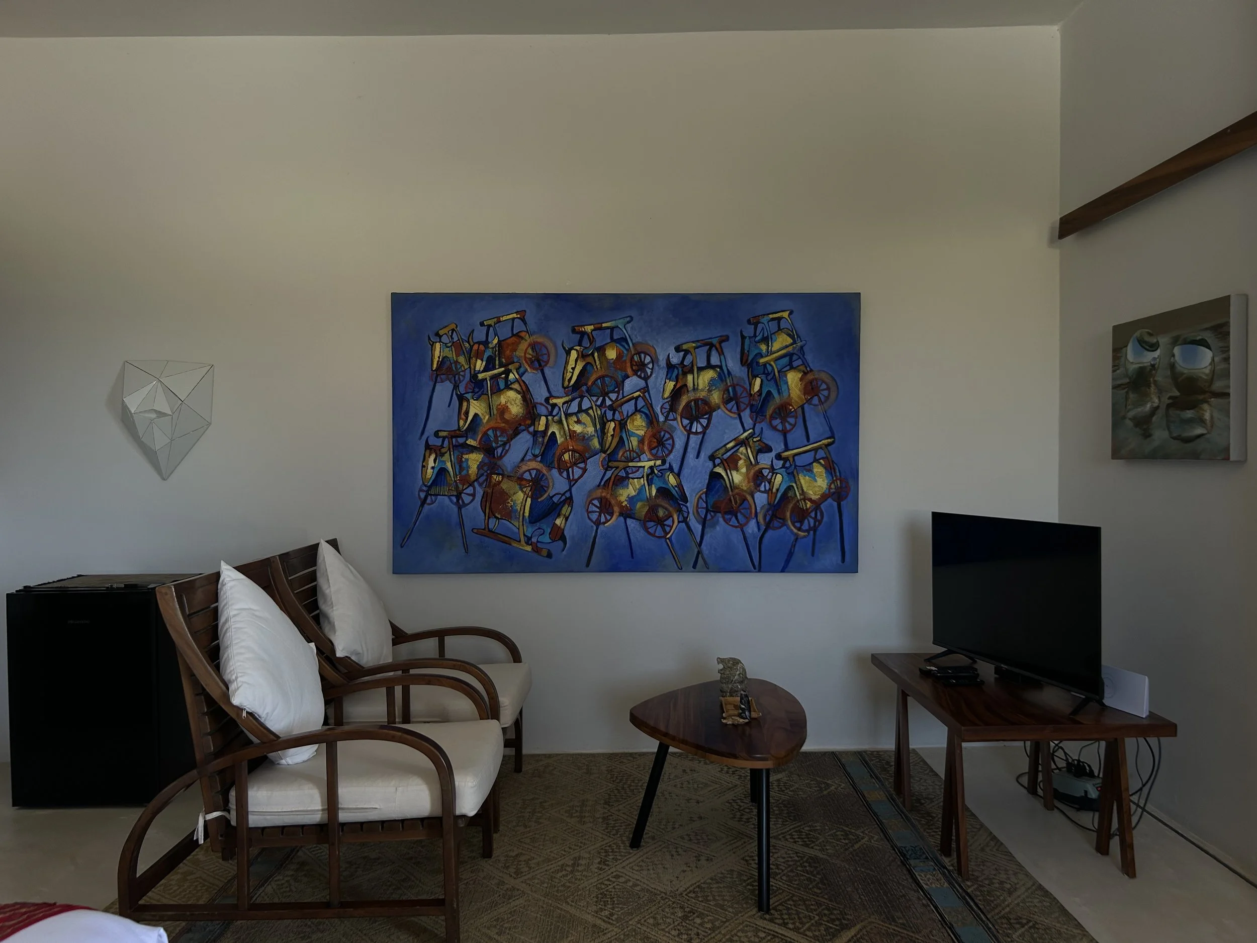 Sala de estar con tres sillones de madera con cojines blancos, una mesa de centro de madera con objetos decorativos, una televisión en un mueble de madera, y varias obras de arte en las paredes, incluyendo un cuadro grande con bicicletas y un espejo 